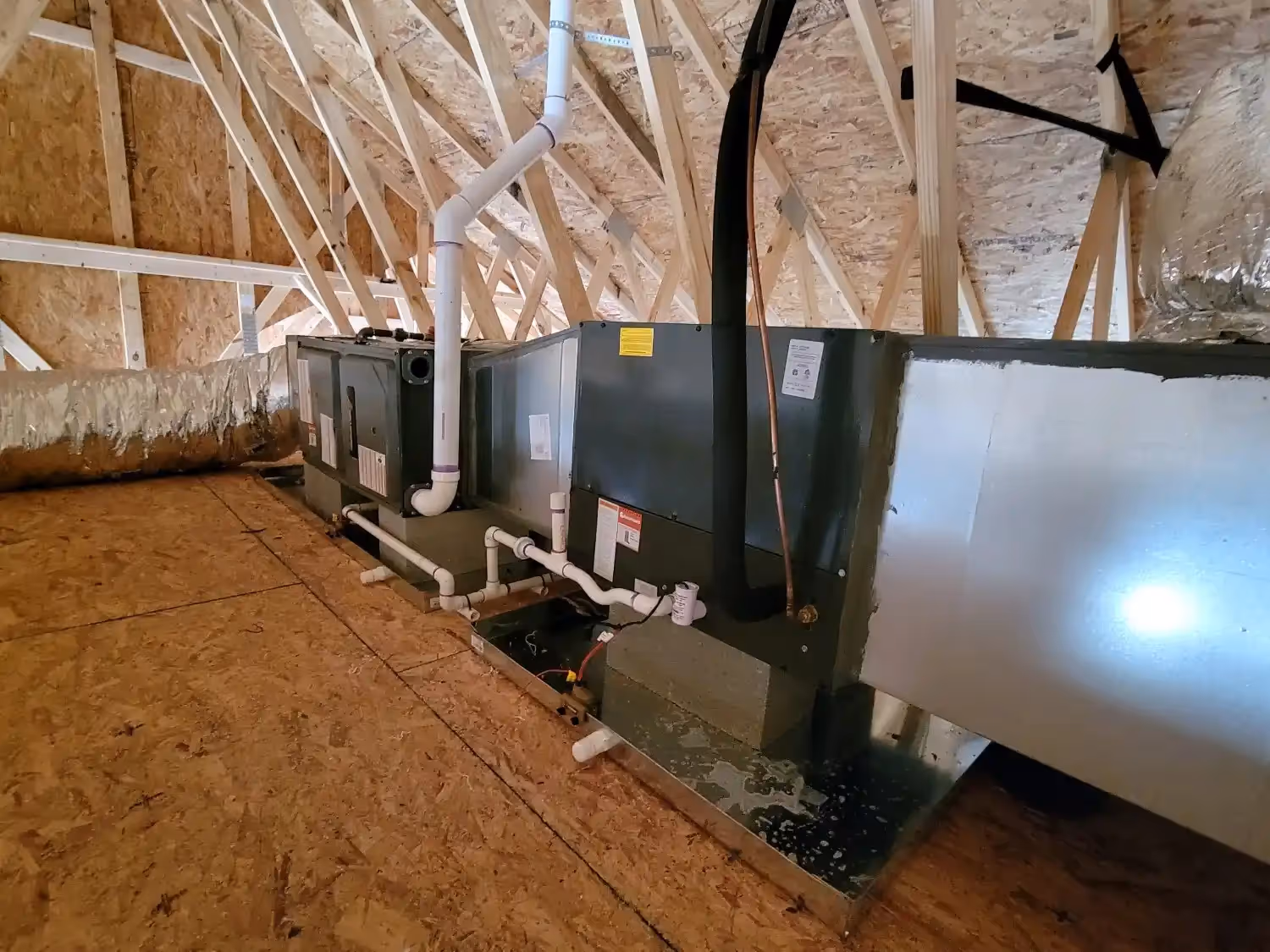 Attic space with two HVAC units, connected to pipes and insulated ducts, on plywood flooring with wooden framing.
