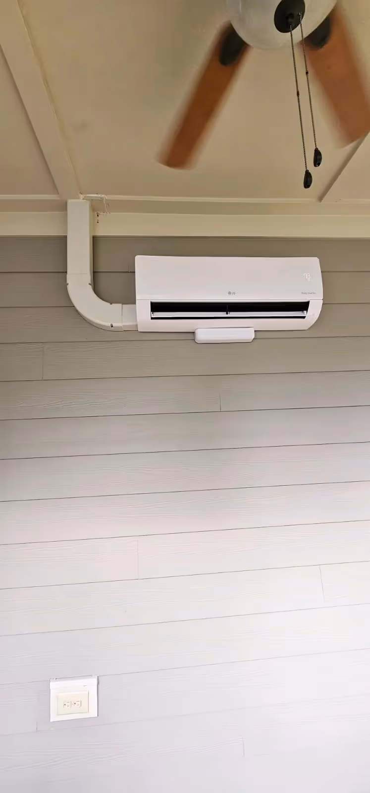 LG air conditioning unit mounted on a wooden-paneled wall, with a connected duct and a ceiling fan above.