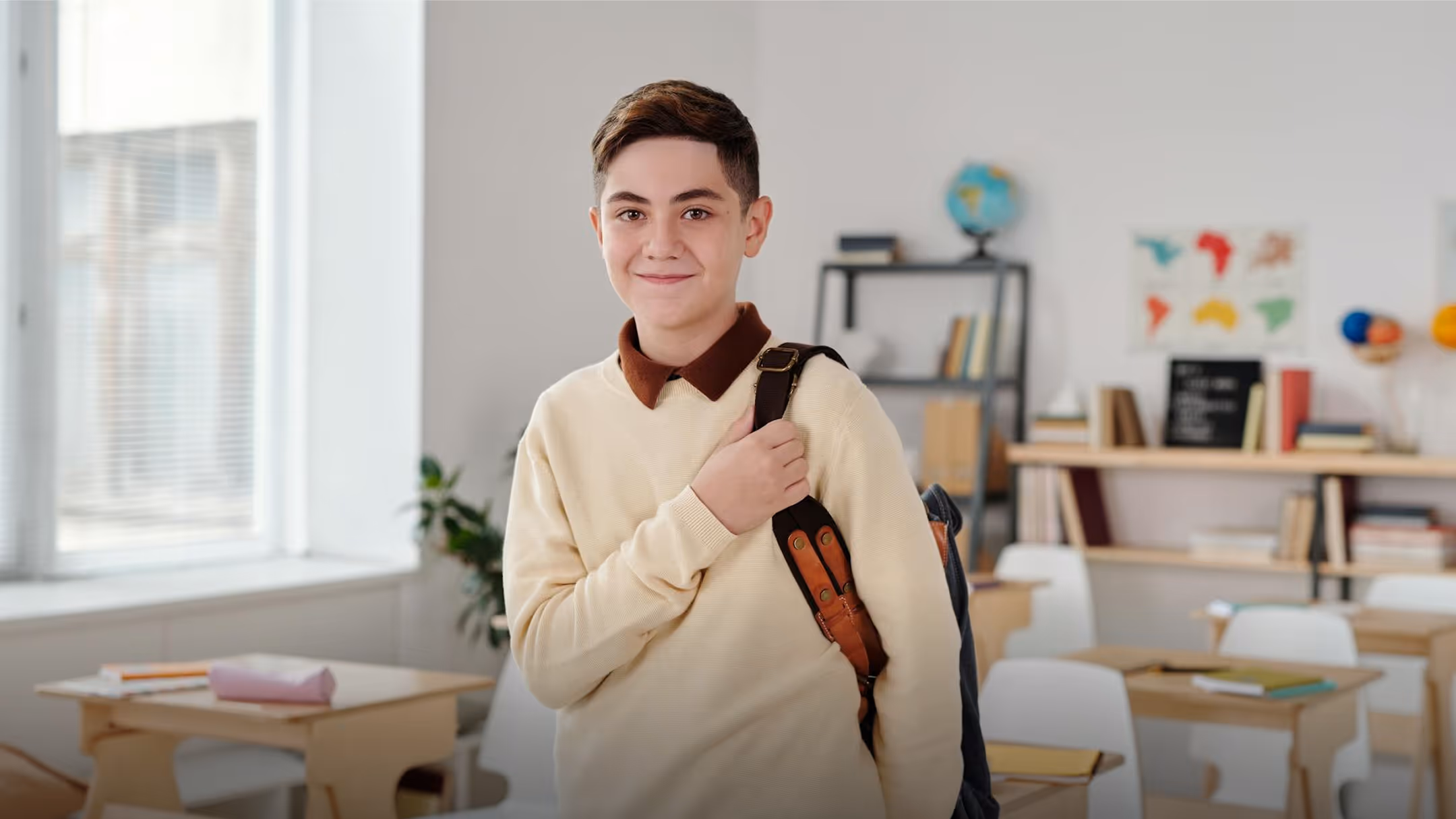 Smiling teenage student with a backpack standing in a classroom with desks, books, and a world map.