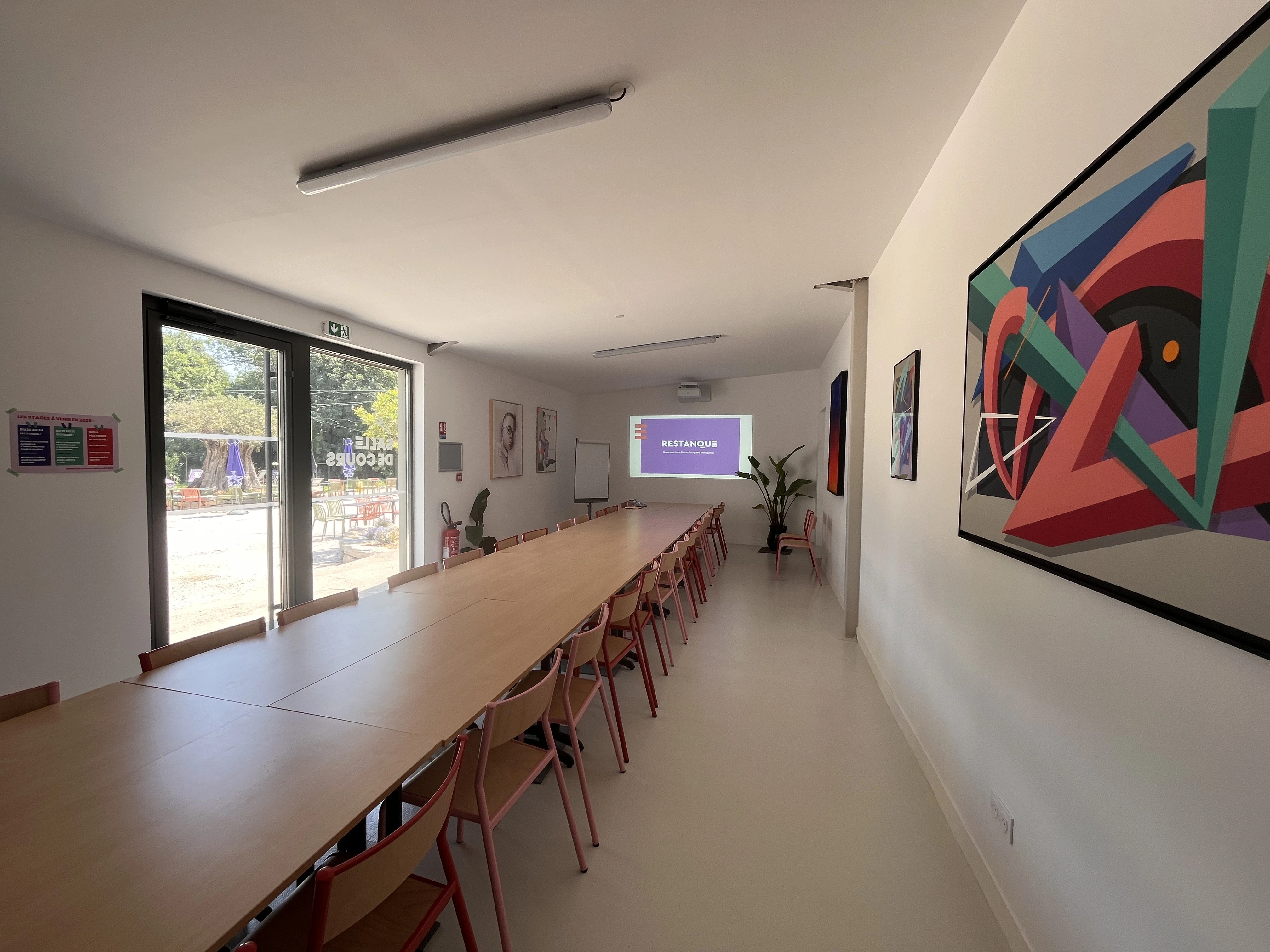 Salle de réunion lumineuse avec une grande table en bois, chaises rouges et vidéoprojecteur affichant le logo Restanque.