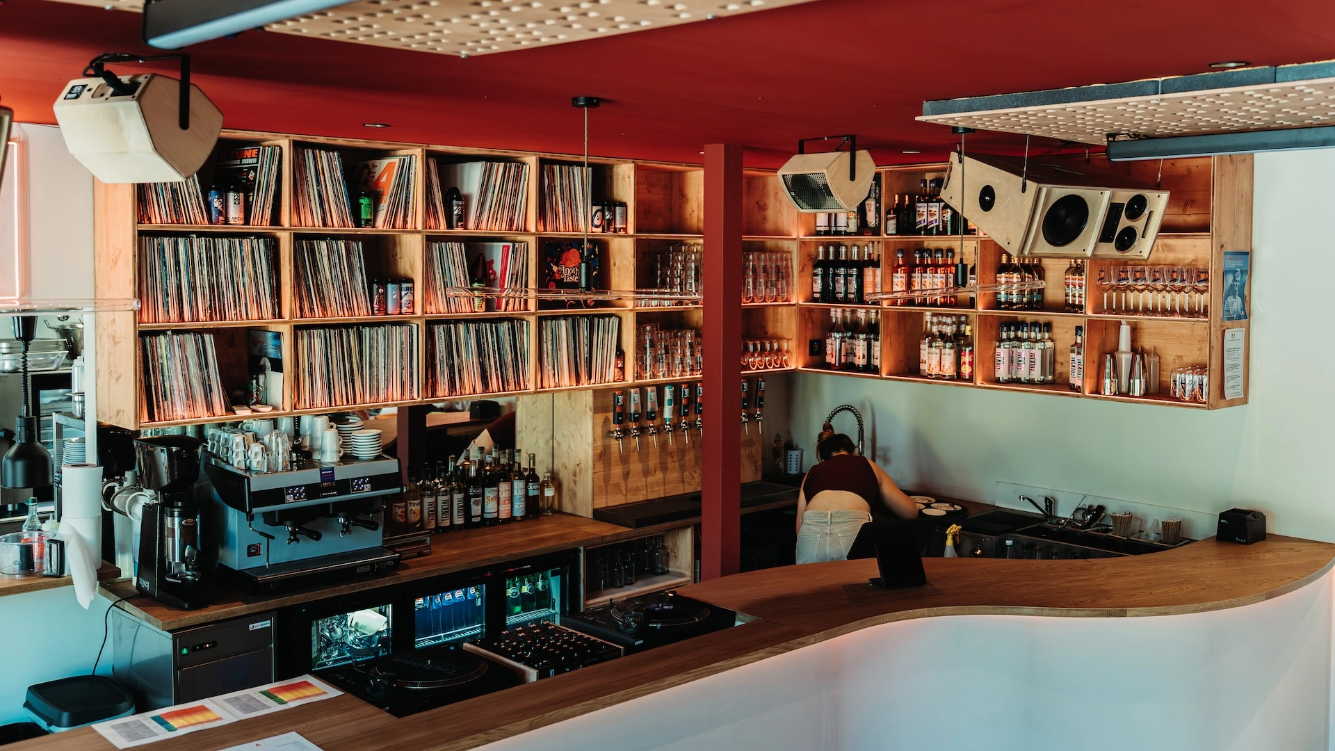 Intérieur d’un bar audiophile avec platines vinyles, étagères remplies de disques et bouteilles, dans une ambiance chaleureuse en bois et rouge.