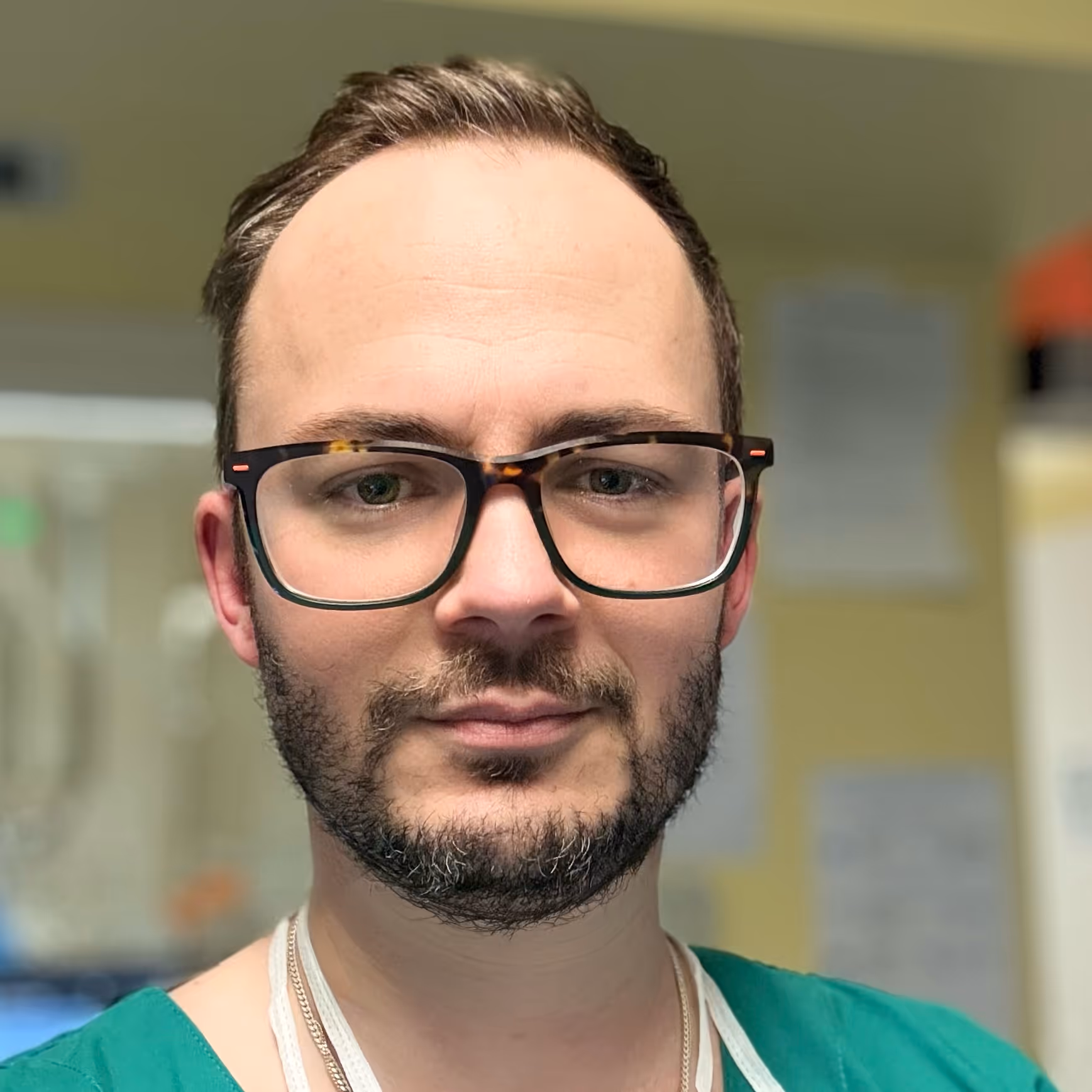 Close-up of a man with glasses, short beard, and green medical scrubs in an indoor setting.