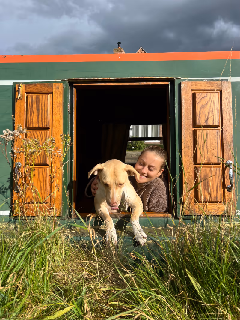 Woman and dog looking out of the hatch of a green narrowboat, smiling.