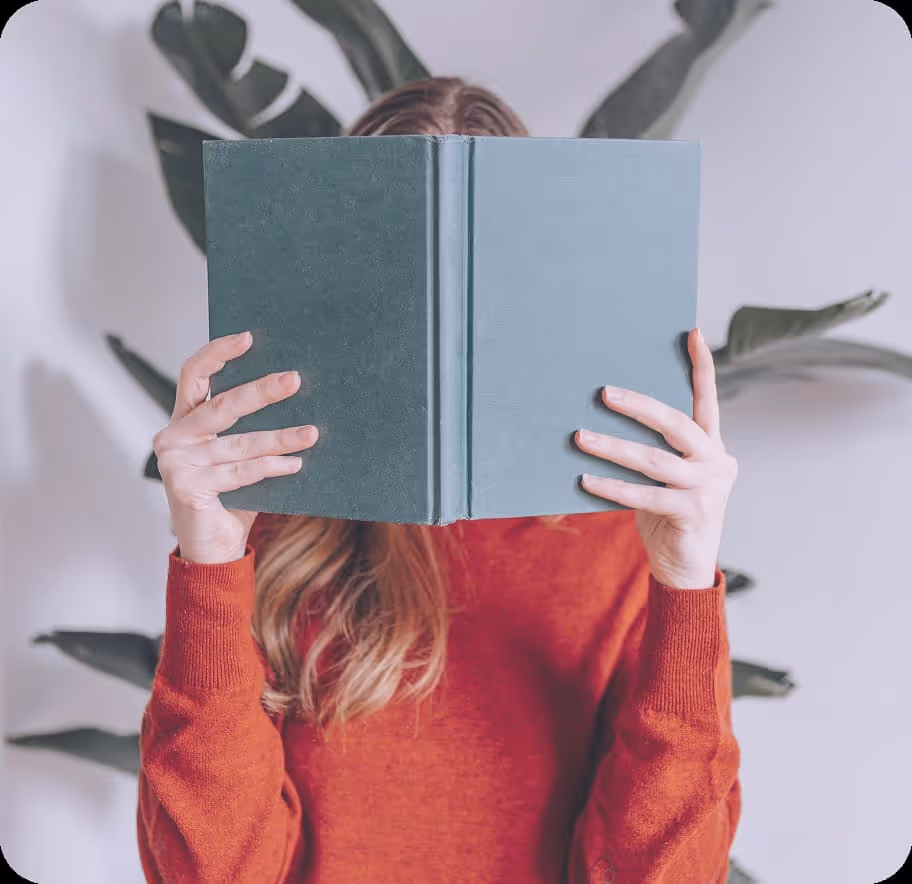 Woman holding a book infront of her face, wearing a red jumper, with a plant behind her