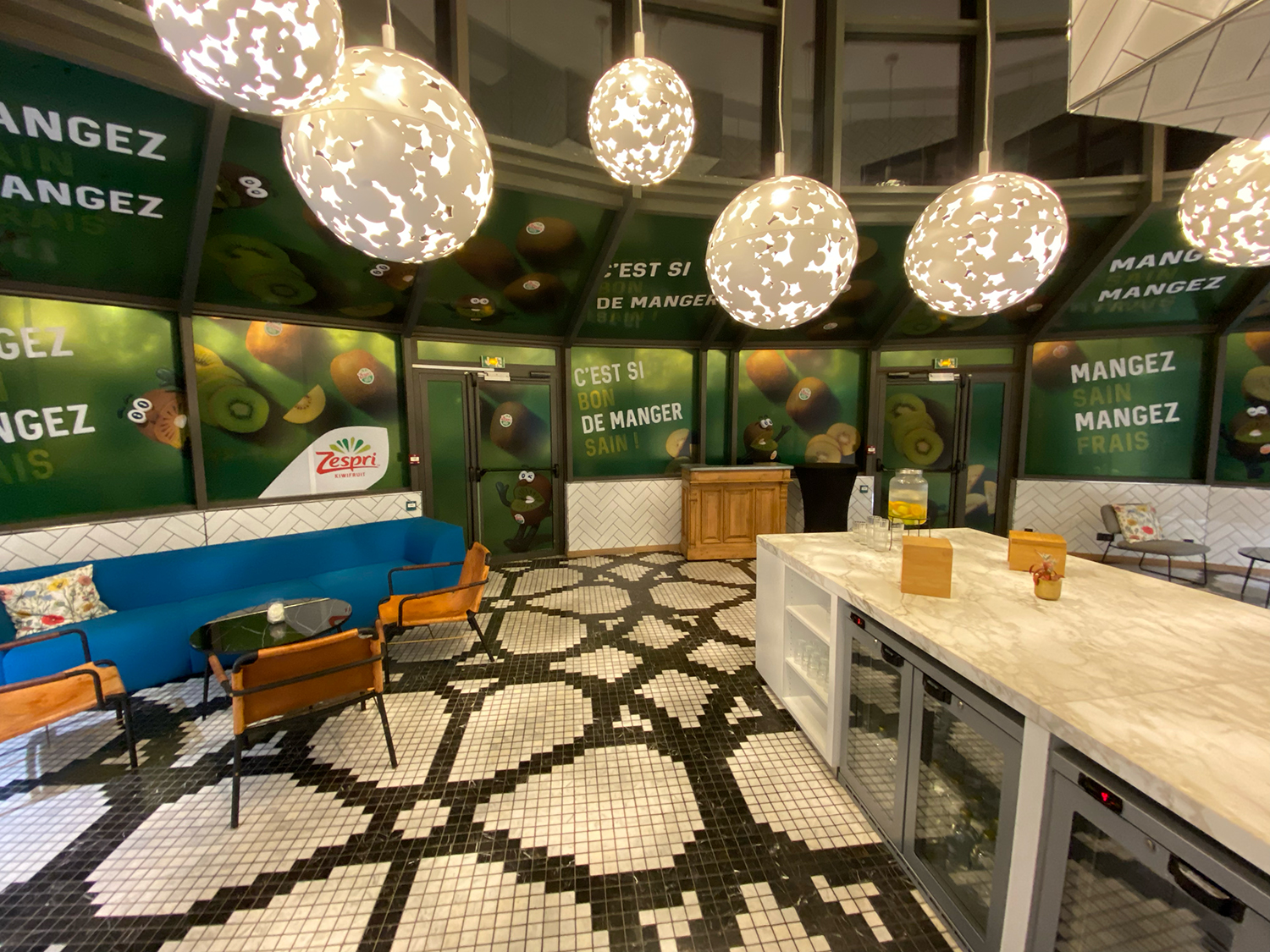 Modern lounge area with a patterned black and white tiled floor, blue sofa, wooden chairs, round table, and a marble countertop with glasses and a water dispenser, surrounded by green walls featuring Zespri kiwi fruit branding and slogans.