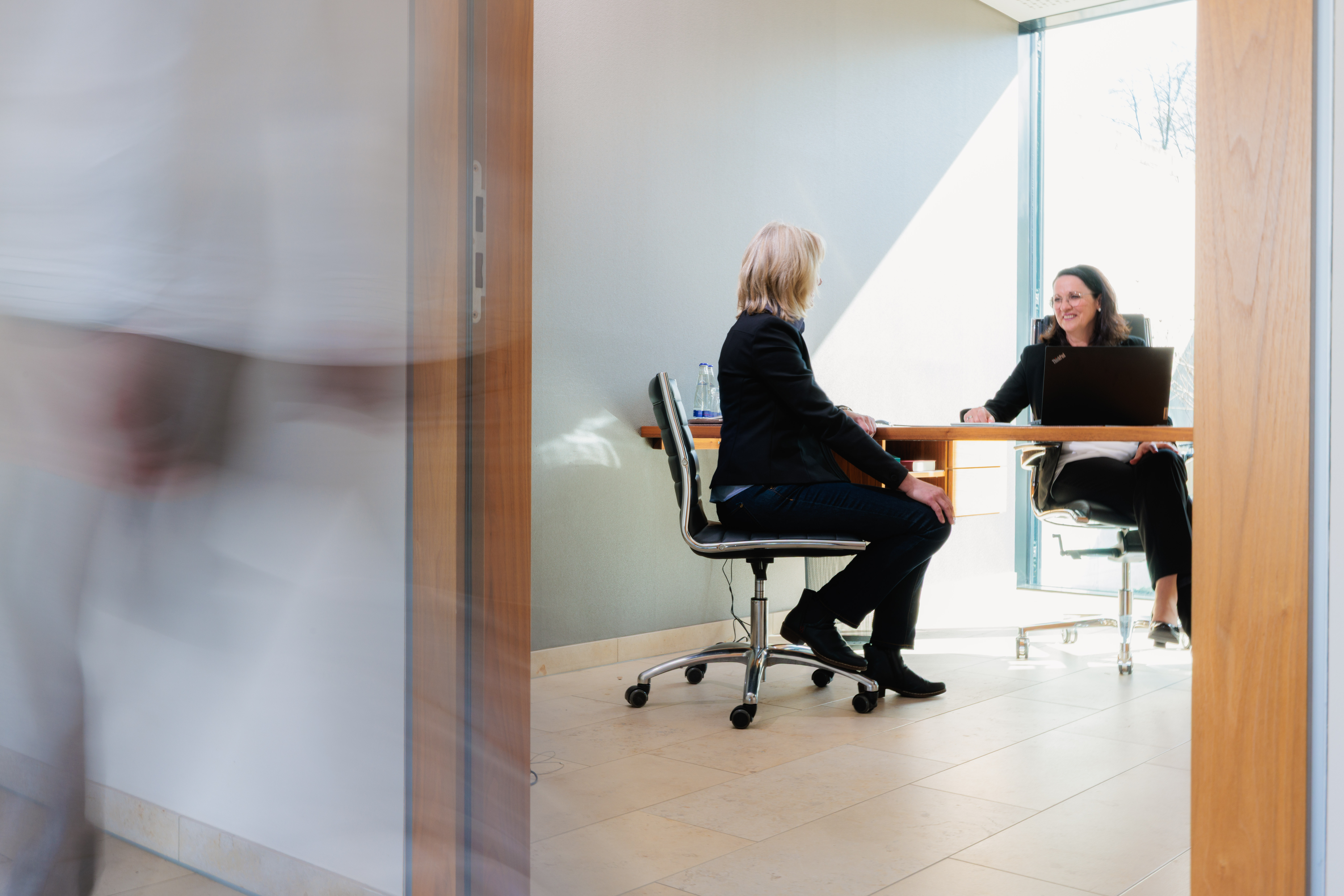 Zwei Frauen im Büro im Gespräch, eine sitzt am Schreibtisch mit Laptop