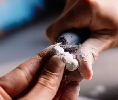 Close-up of hands shaping a white clay figurine with a small rotary tool.