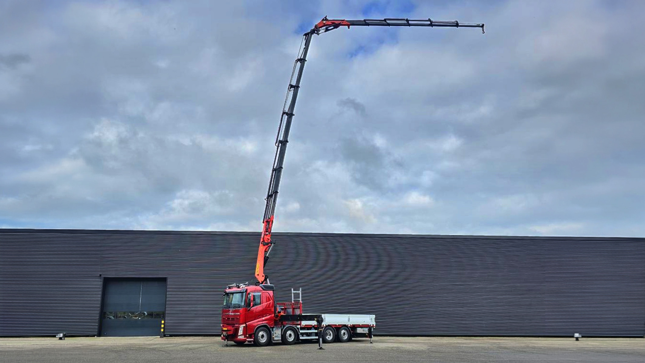 Roter Lastwagen mit ausgestrecktem Kranarm vor einer großen grauen Industriehalle unter bewölktem Himmel.