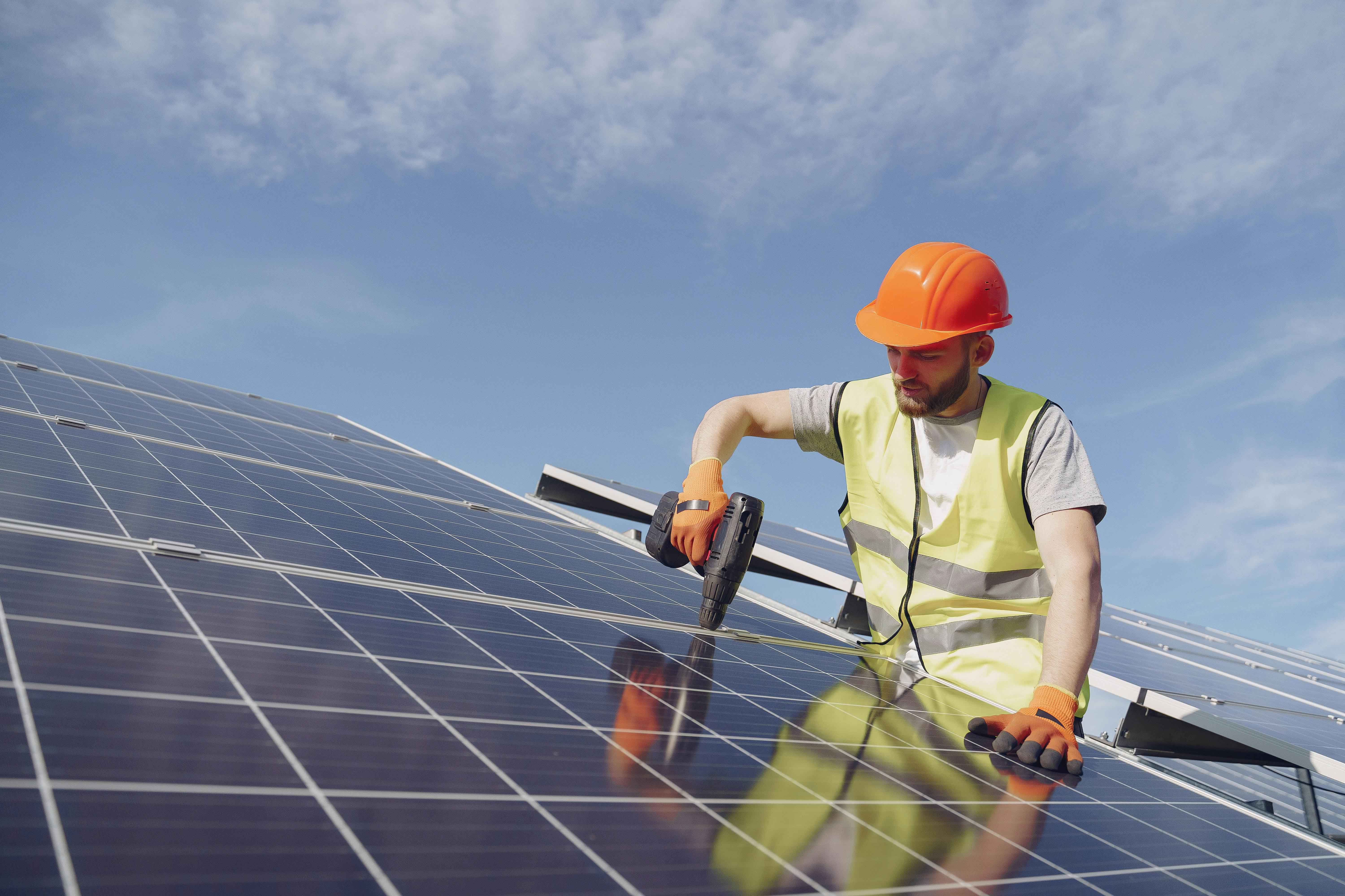 Ein Arbeiter mit orangefarbenem Schutzhelm und gelber Warnweste installiert Solarpaneele mit einem Akkuschrauber unter blauem Himmel.