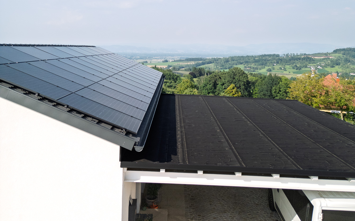 Haus mit Solarpaneelen auf dem geneigten Dach und flachem schwarzen Carportdach, im Hintergrund grüne Landschaft mit Bäumen und Hügeln.