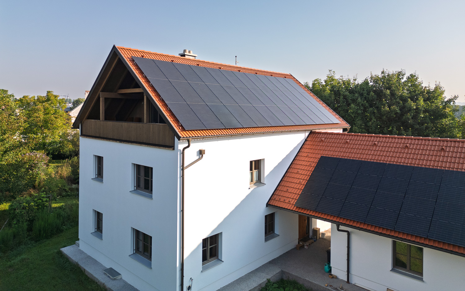 Modernes weißes Haus mit roten Dachziegeln und Solarpanels auf den Dächern, umgeben von Bäumen.