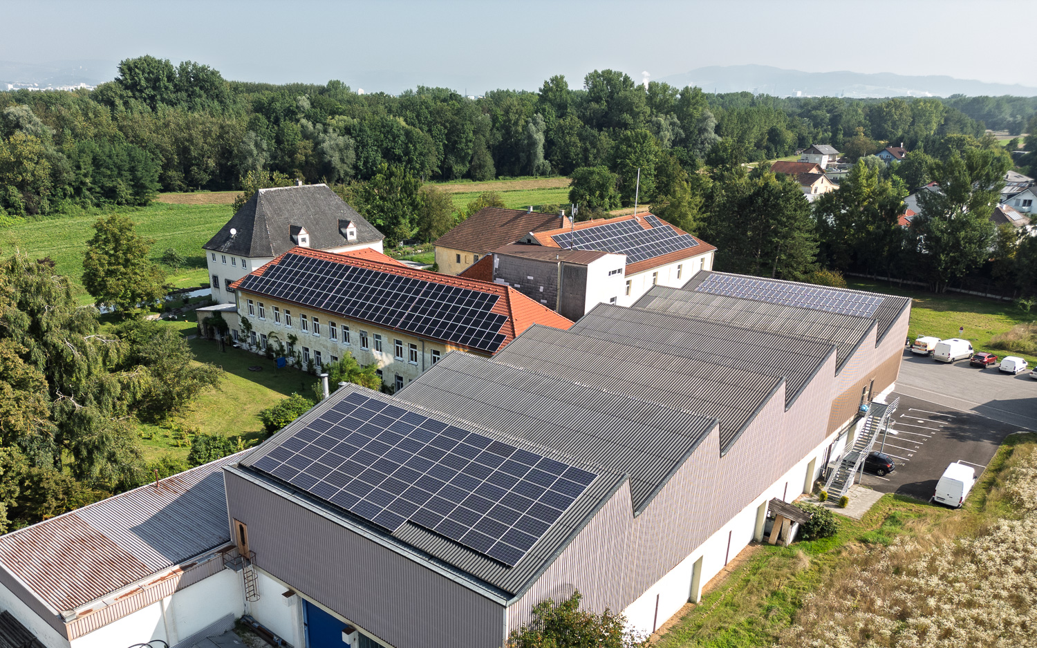 Luftaufnahme von Gebäuden mit Solarpaneelen auf den Dächern, umgeben von Bäumen und Feldern.
