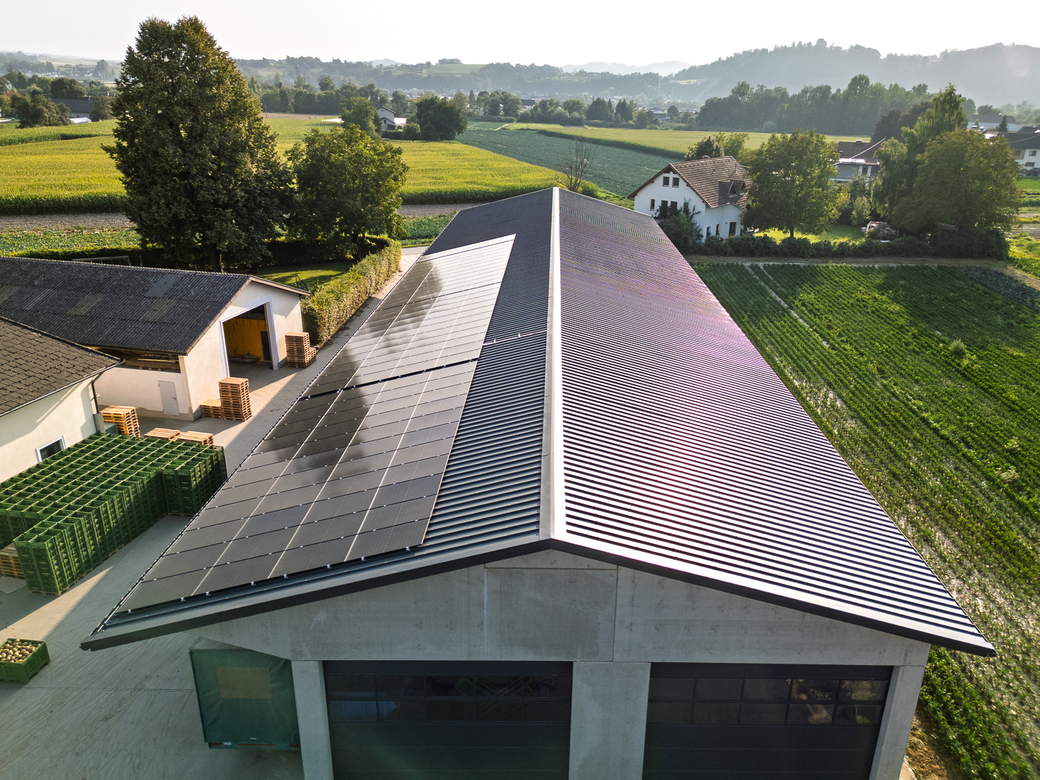 Landwirtschaftliches Gebäude mit Solarpanelen auf dem Dach, umgeben von grünen Feldern und Bäumen.