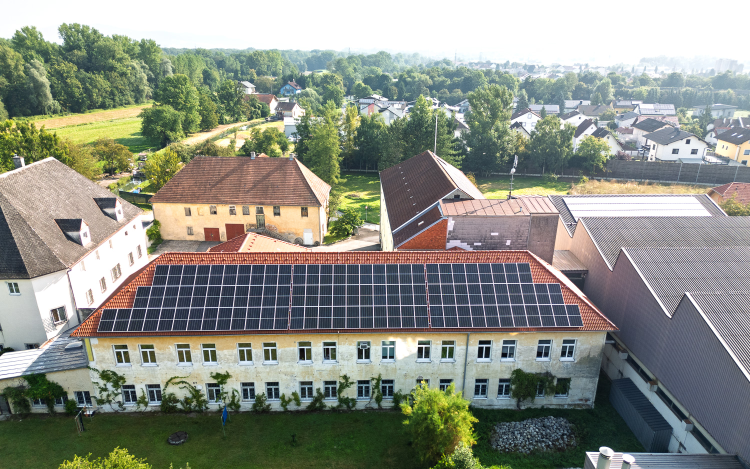 Luftaufnahme eines älteren Gebäudes mit Solarpaneelen auf dem Dach, umgeben von weiteren Gebäuden und viel Grün.