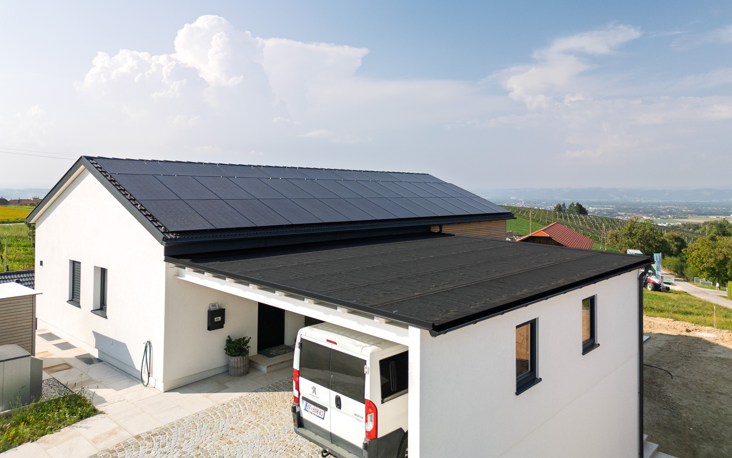 Modernes weißes Haus mit Flachdach-Carport und Solarpaneelen auf dem Satteldach, umgeben von ländlicher Landschaft.