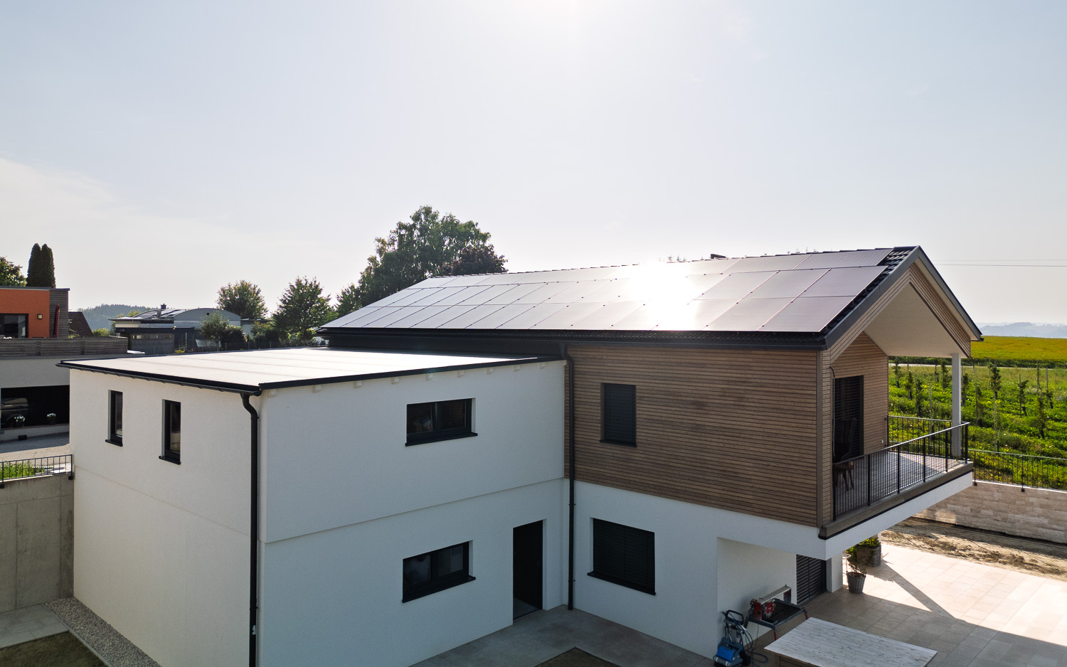 Modernes Einfamilienhaus mit weißer Fassade, Holzverkleidung und Solarpaneelen auf dem Dach bei sonnigem Himmel.