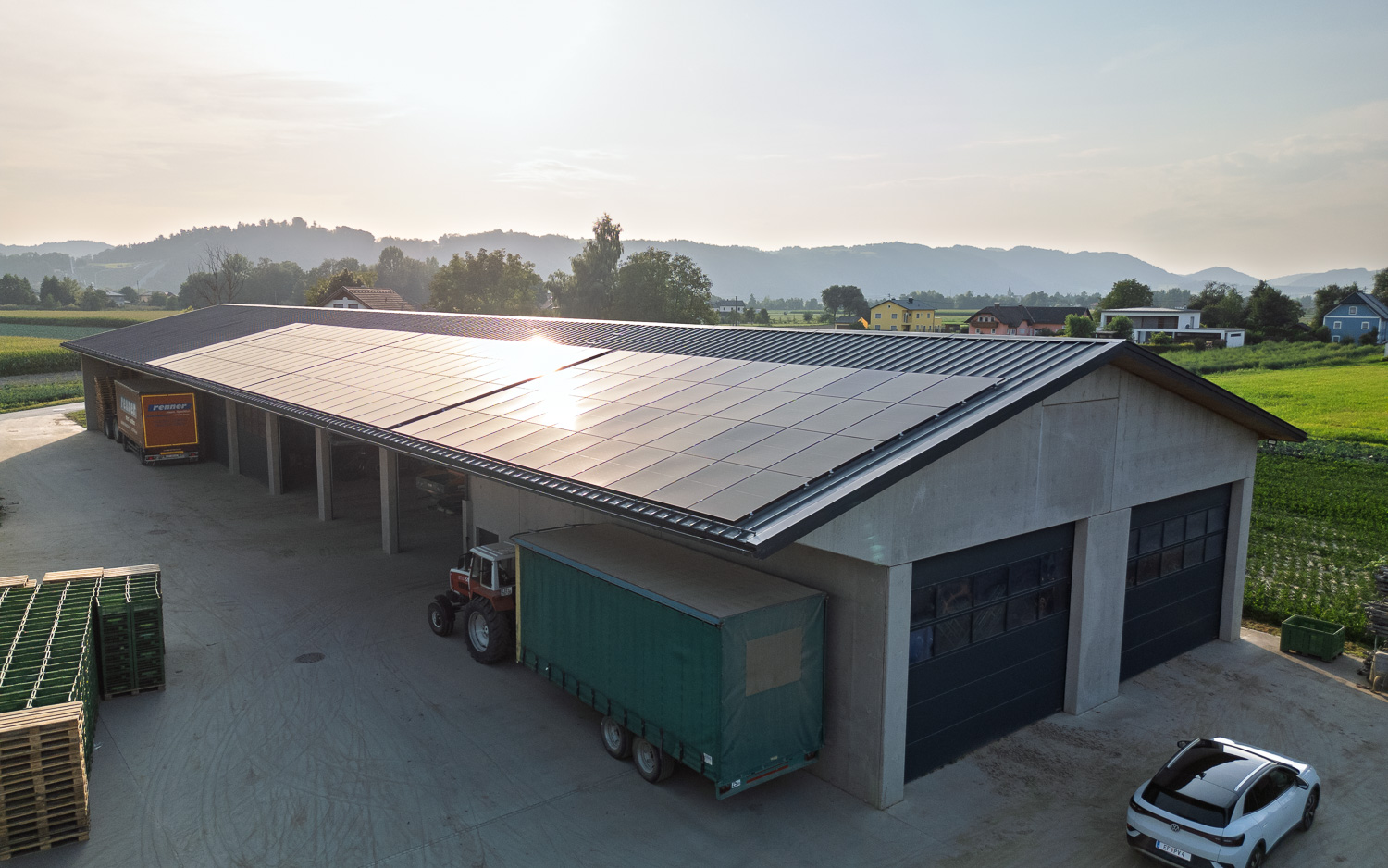 Landwirtschaftliches Gebäude mit einer Dachfläche voller Solarpanels, umgeben von offenem Land und einzelnen Fahrzeugen.