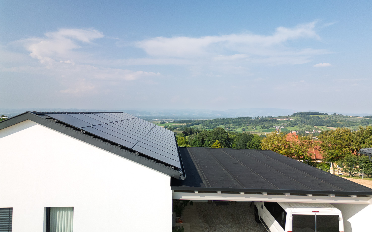 Haus mit Solarpaneelen auf dem schrägen Dach und Carport mit weißem Fahrzeug vor einer grünen Hügellandschaft.