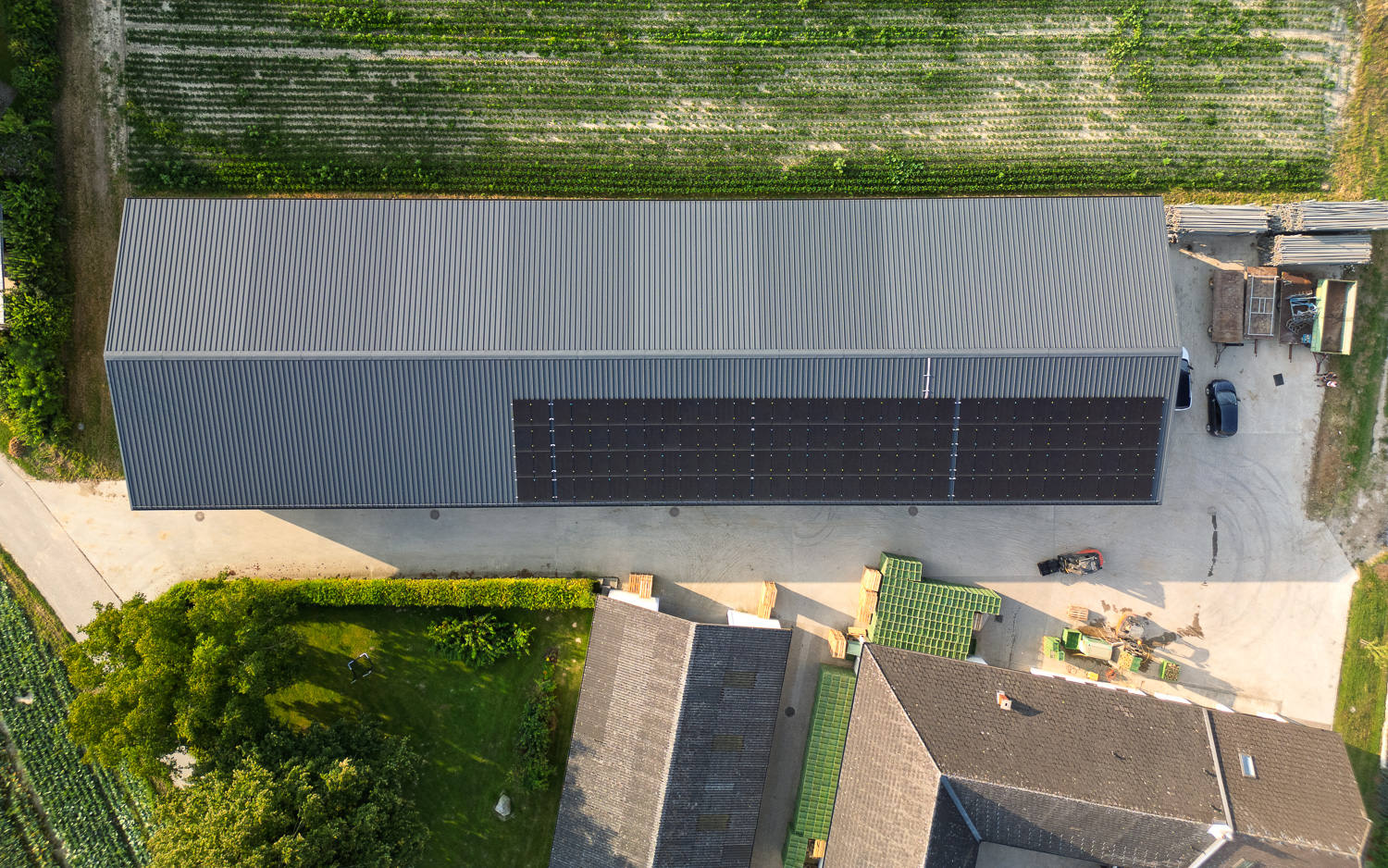 Luftaufnahme eines großen Gebäudes mit Solarpaneelen auf dem Dach, umgeben von Feldern und angrenzenden kleineren Dächern.