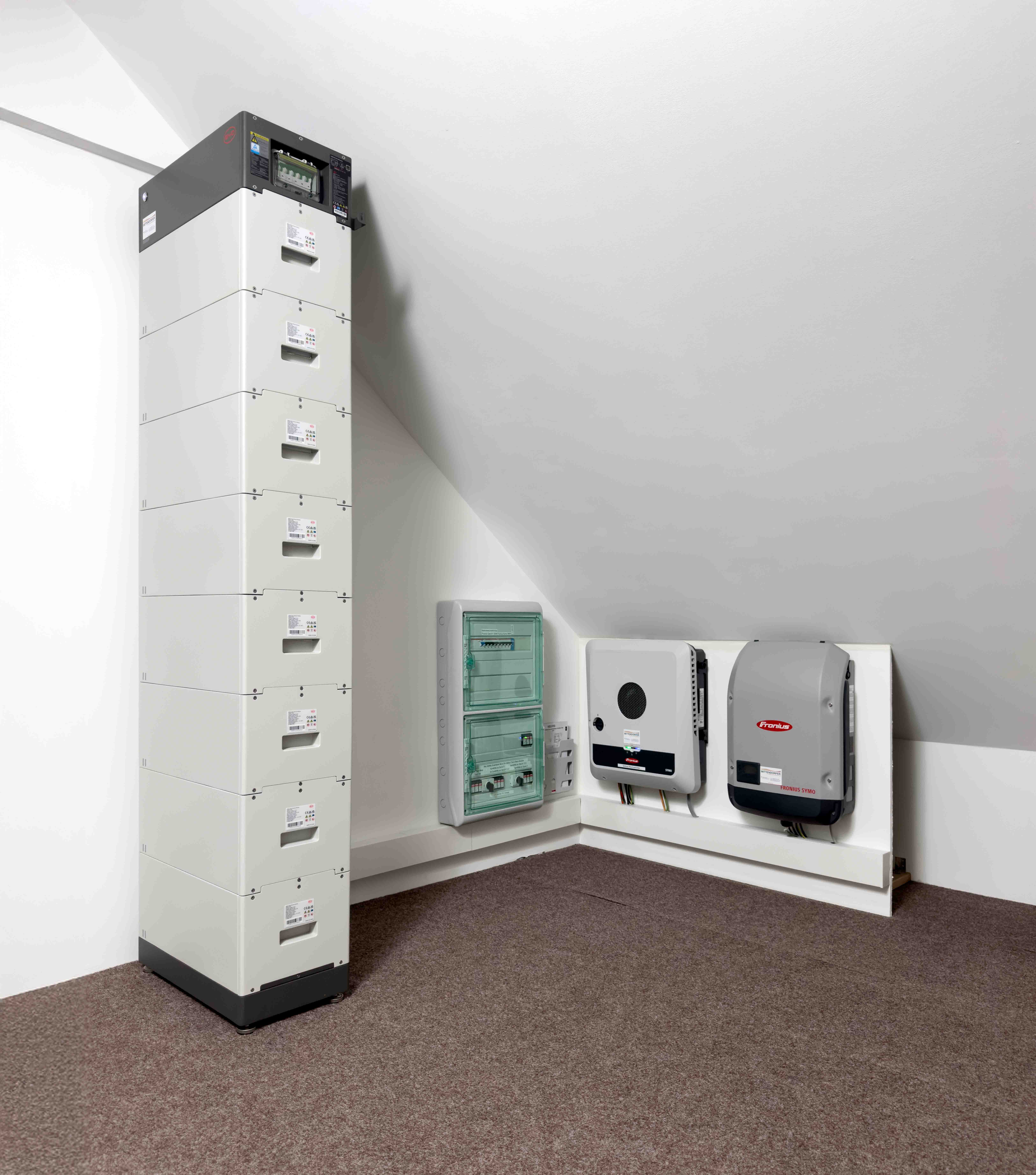 Starker Energiespeicher mit mehreren weißen Boxmodulen und zwei Fronius Wechselrichtern in einem Raum mit schräger Decke.