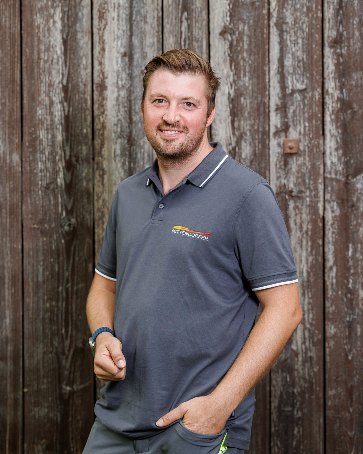 Mann mit kurzem blonden Haar und Bart, trägt ein graues Poloshirt mit ‚MITTENDORFER‘-Logo, steht vor einer hölzernen Wand und lächelt in die Kamera.