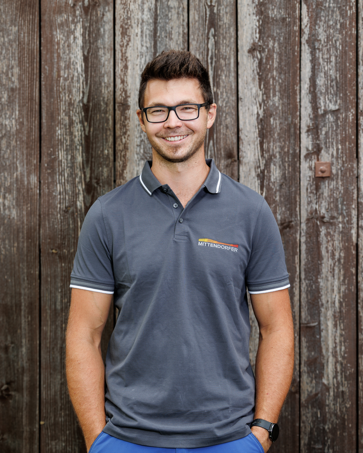 Junger Mann mit Brille und grauem Poloshirt mit Mittendorfer-Logo vor hölzernem Hintergrund lächelt.
