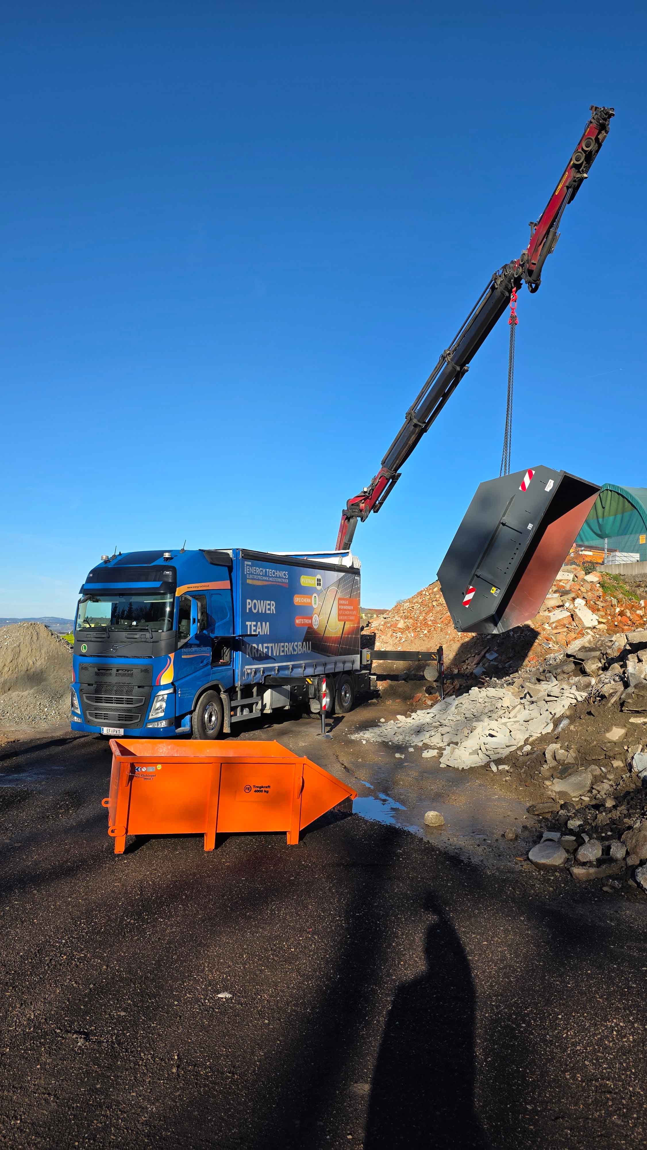 Blauer Lastwagen mit Aufdruck 'Power Team Kraftwerksbau' hebt mit Kran eine graue Mulde an einer Baustelle.