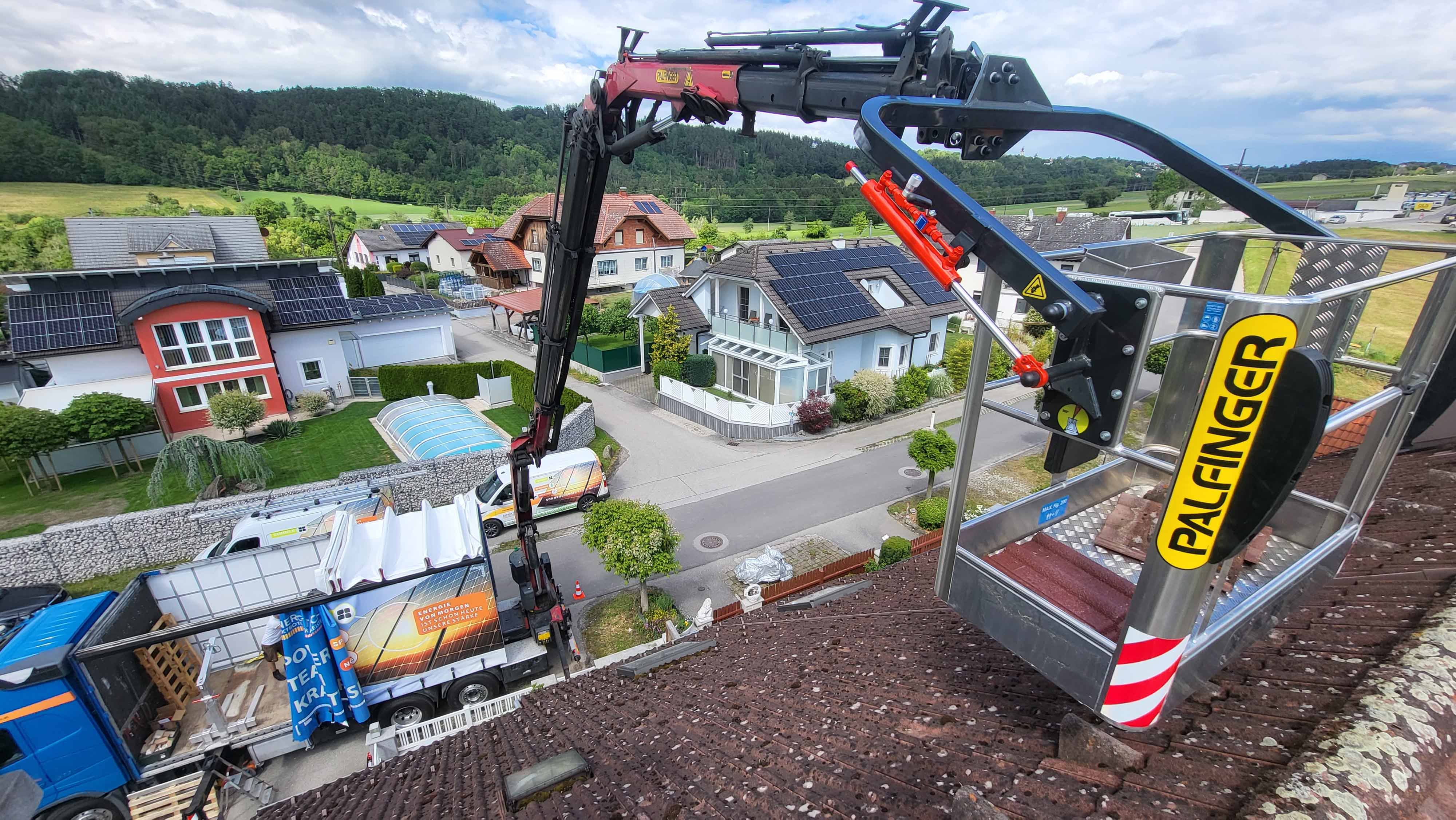Hebekorb mit Palfinger-Logo an einem Kran über einem LKW und Wohnhäusern mit Solarpanels auf den Dächern in ländlicher Umgebung.
