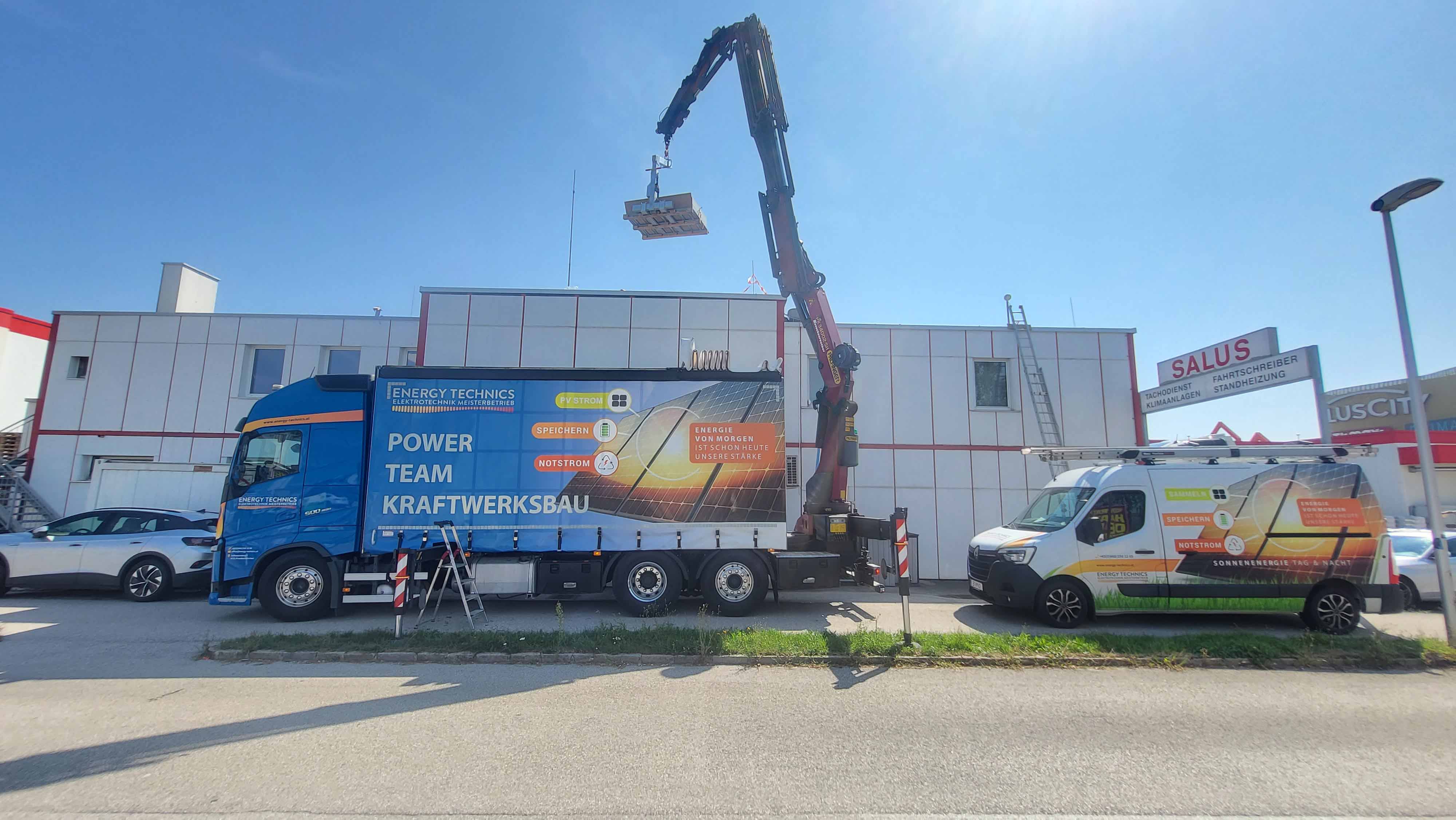 Lastwagen mit Kran hebt eine Palette vor einem Gebäude, daneben ein weißer Transporter mit Solarpanel-Grafik und Werbetext für erneuerbare Energie.