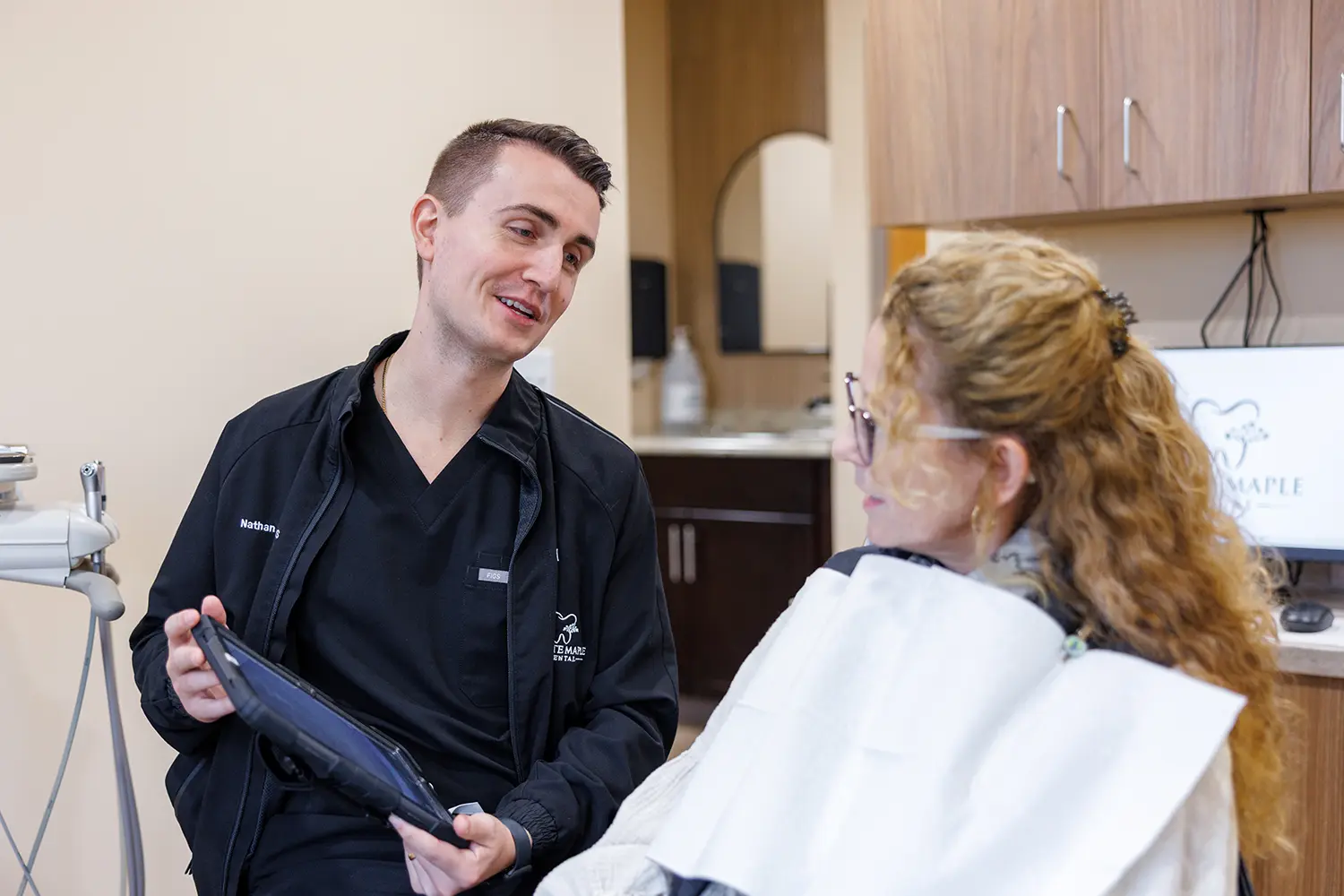 A male dentist wearing black scrubs and a jacket holds a tablet and talks to a female patient sitting in a dental chair with a white bib, in a dental office.