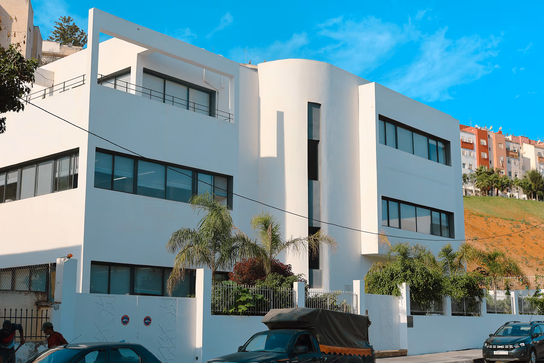 Modern white multi-story building with large windows, surrounded by palm trees and parked cars under a clear blue sky.