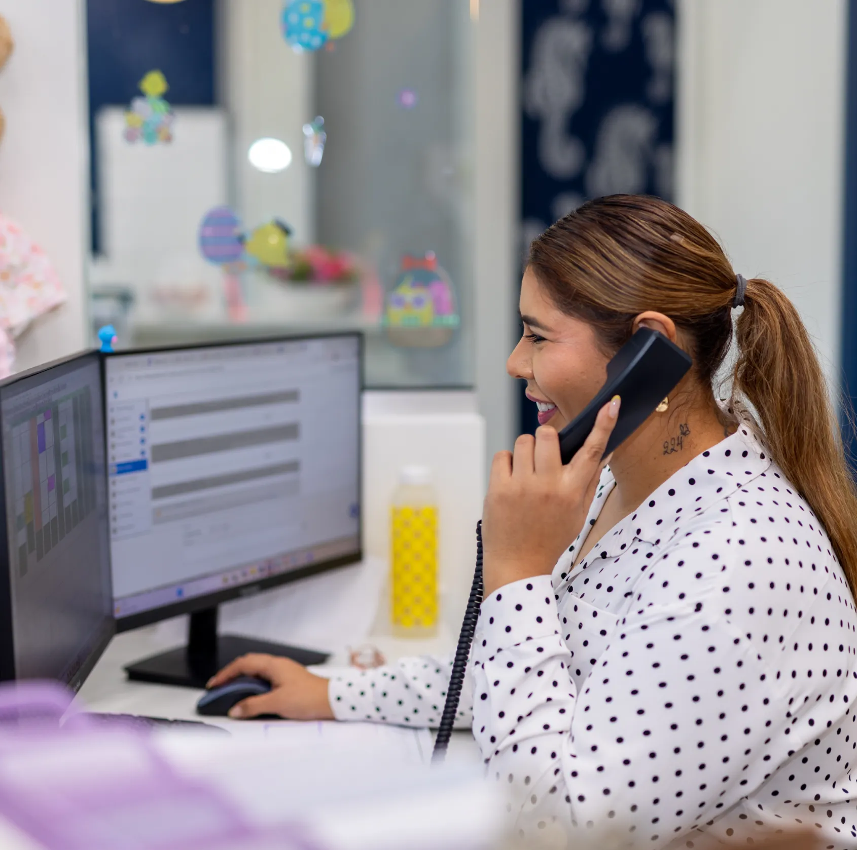 Woman in a white polka dot shirt smiling and talking on a phone while using a computer with dual monitors at a desk.
