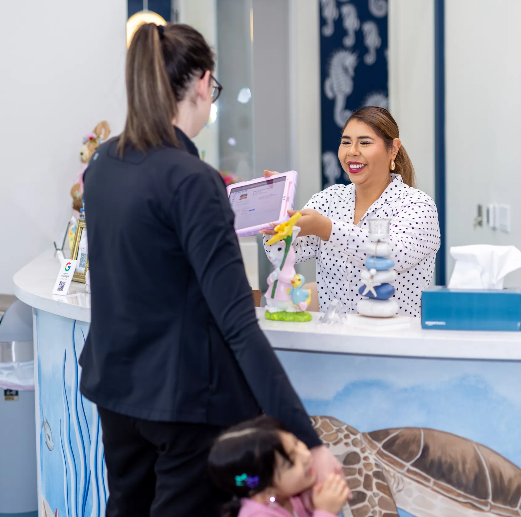 Smiling receptionist in a polka-dot blouse showing information on a tablet to a woman holding a young child at a sea turtle-themed reception desk.