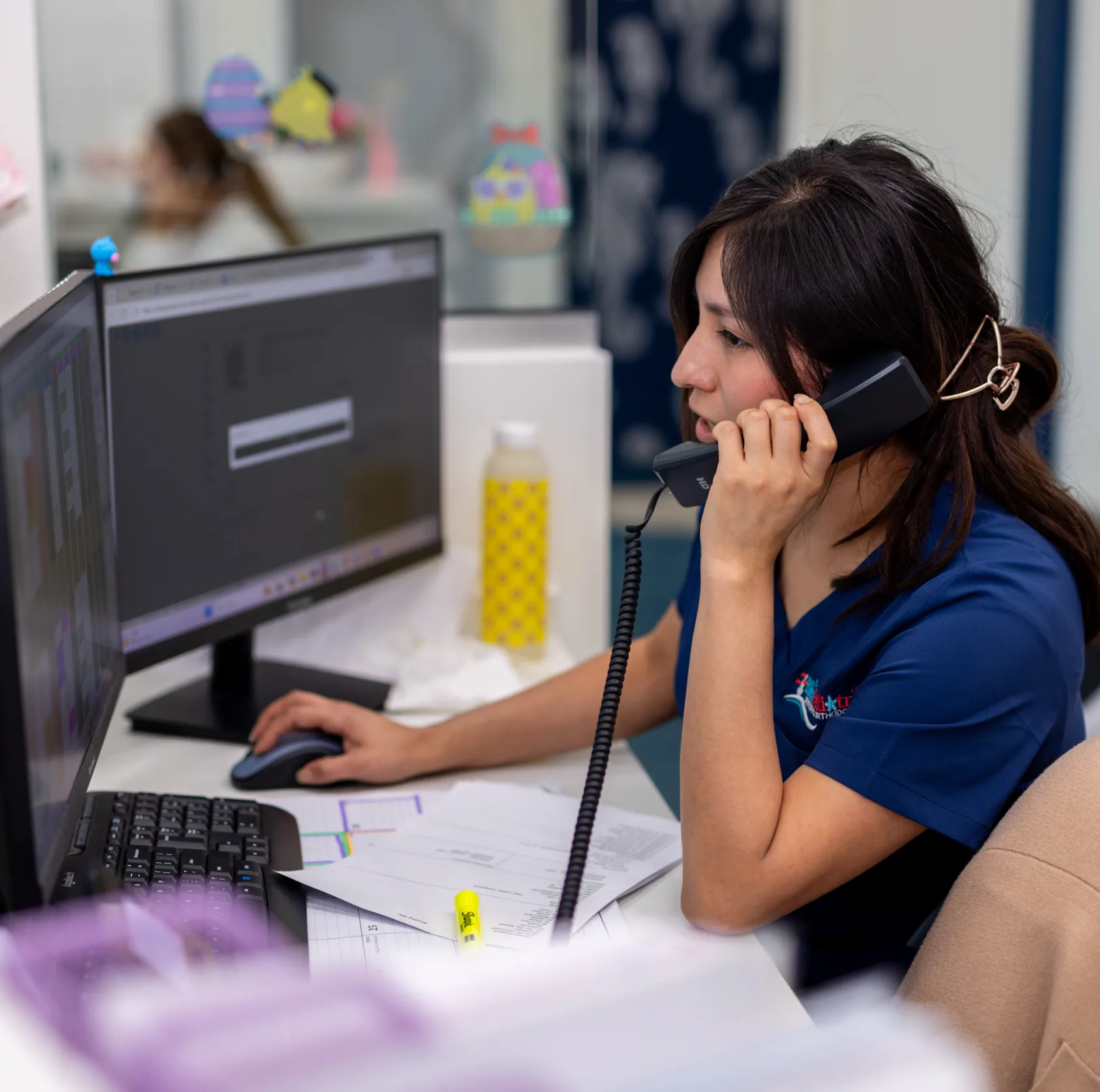 Woman in blue shirt talking on a corded phone while using a computer mouse at a desk with papers and two monitors.