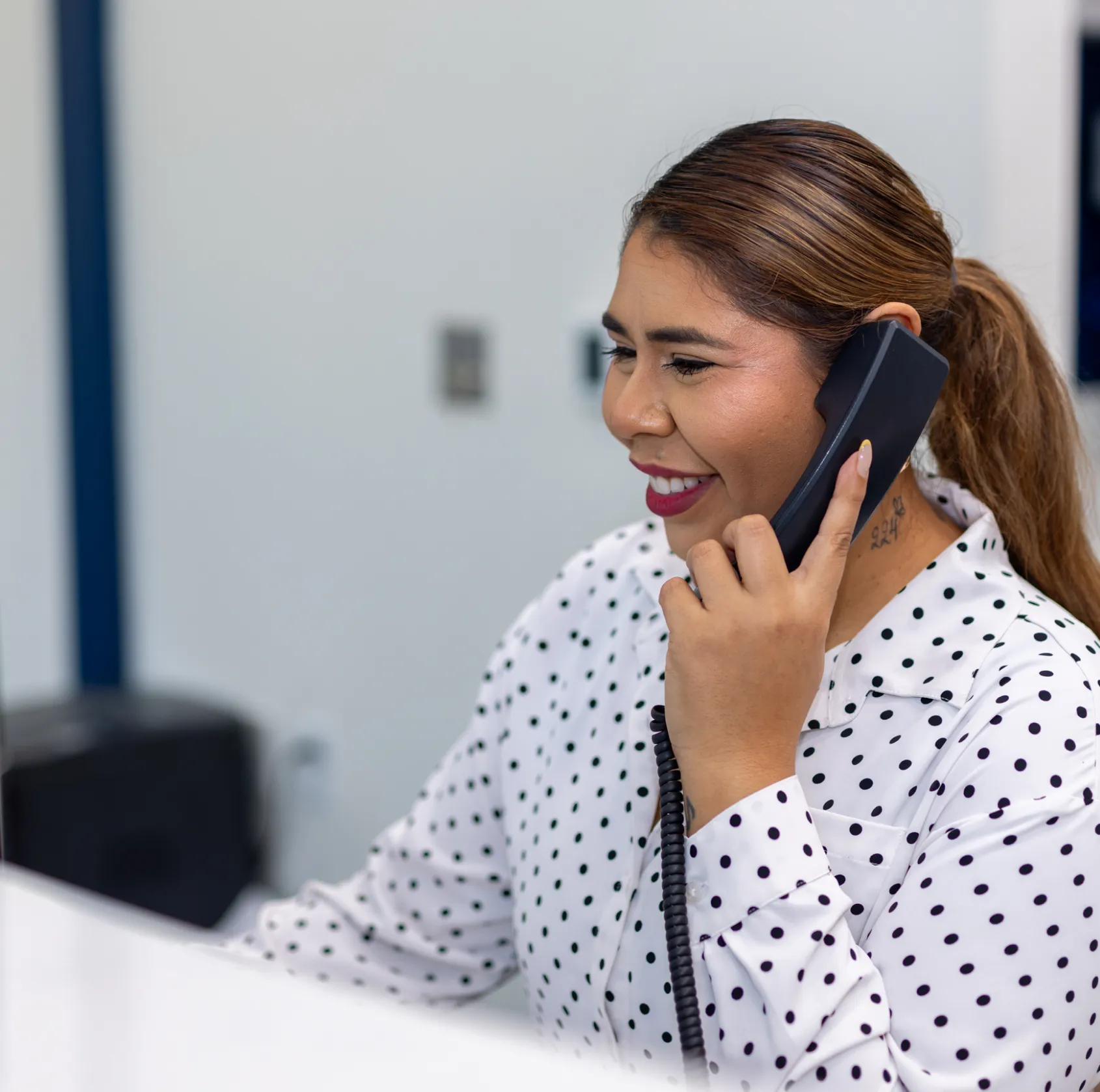 Smiling woman with a ponytail and polka dot blouse talking on a black corded telephone in an office setting.