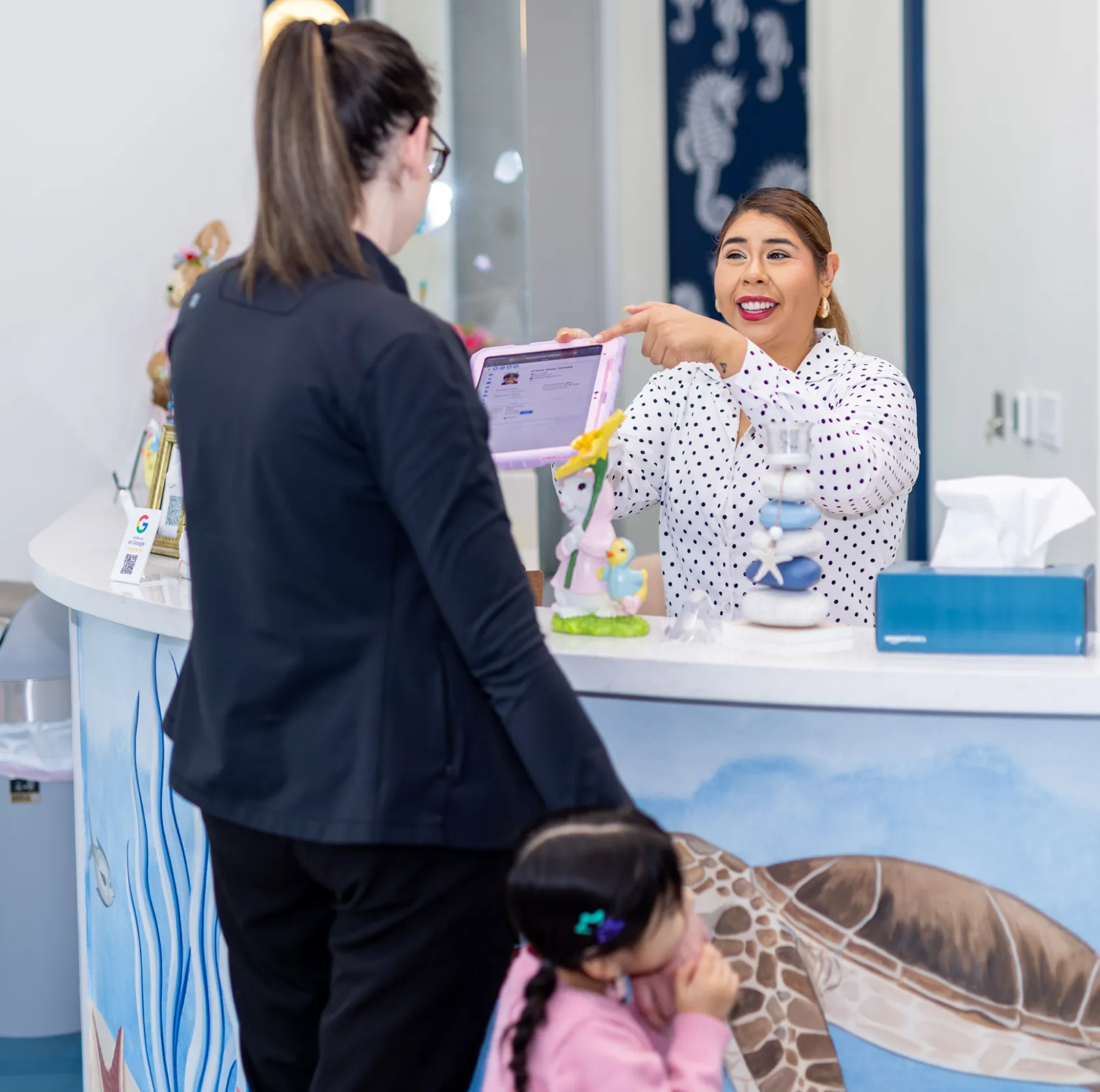 Smiling receptionist in a white polka dot shirt holding a tablet and pointing at its screen while talking to a standing woman with a child at the reception desk with sea turtle artwork.