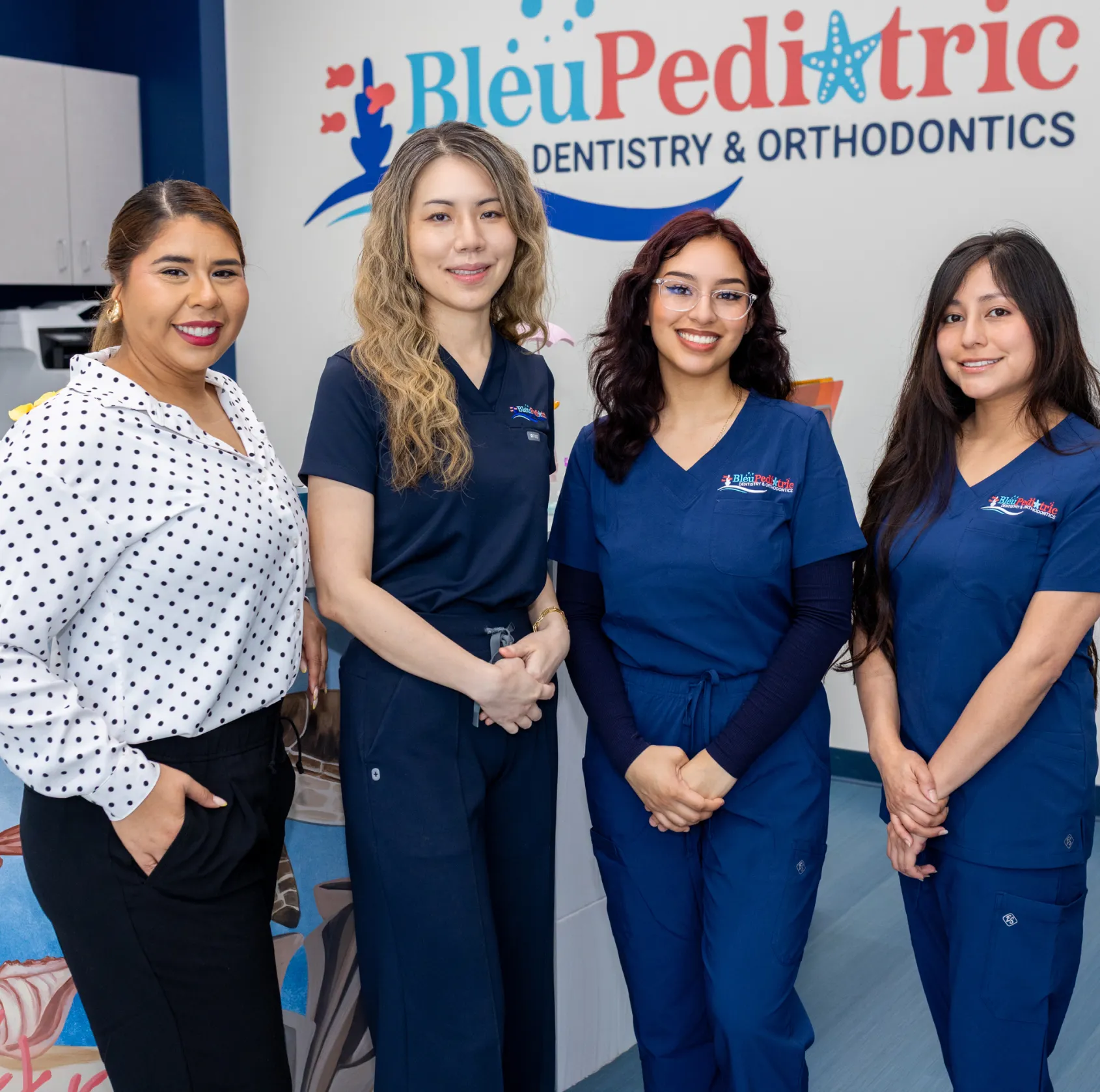 Four smiling female dental professionals standing in front of a Bleu Pediatric Dentistry & Orthodontics sign.
