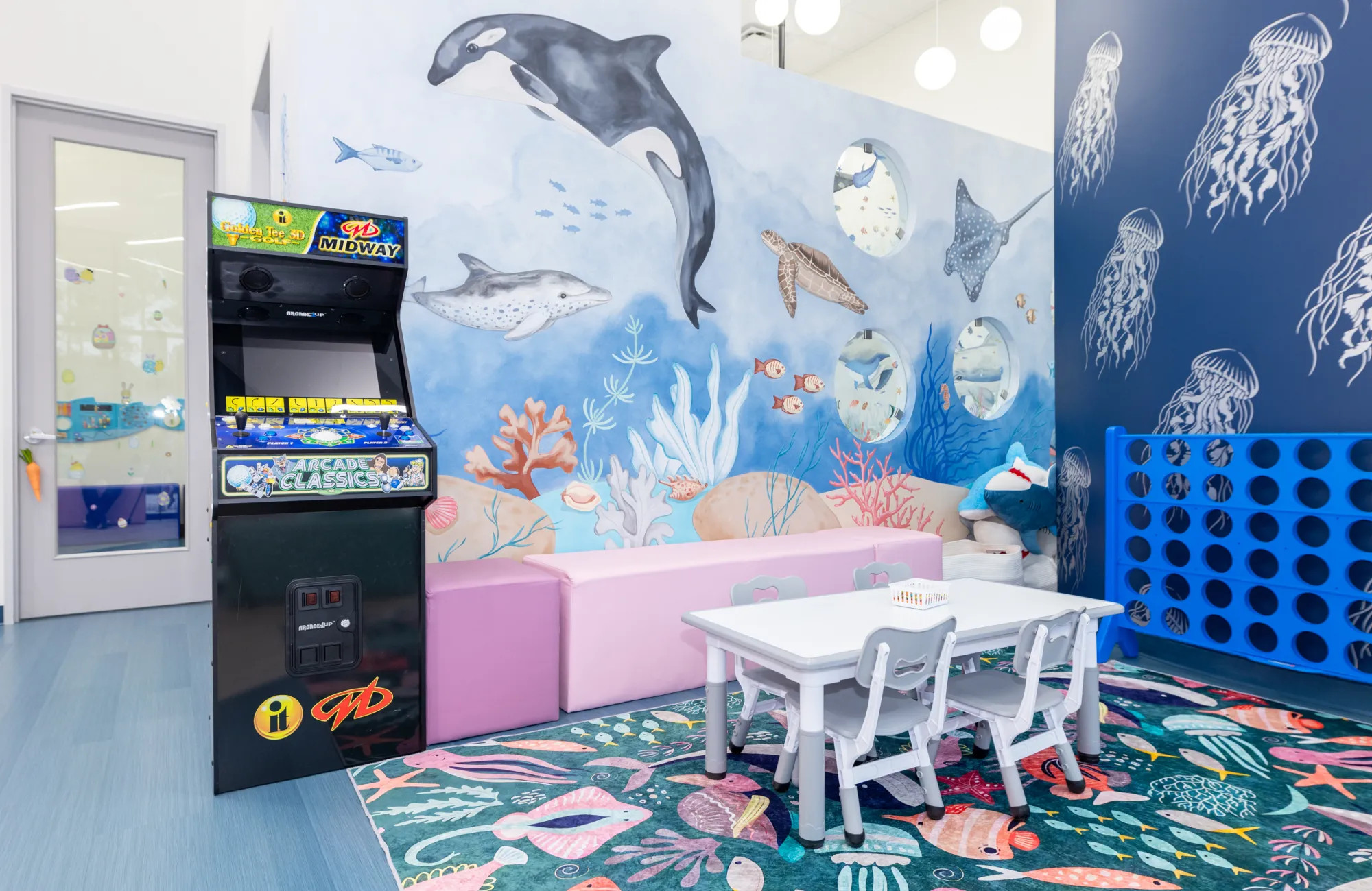 Children's playroom with ocean-themed mural featuring whales, dolphins, and turtles, a classic arcade game, small table with chairs, and large Connect Four game on a colorful fish-patterned rug.