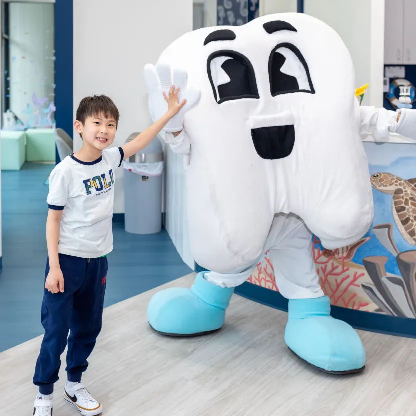 Boy smiling and giving a high five to a person in a large tooth mascot costume inside a dental clinic.