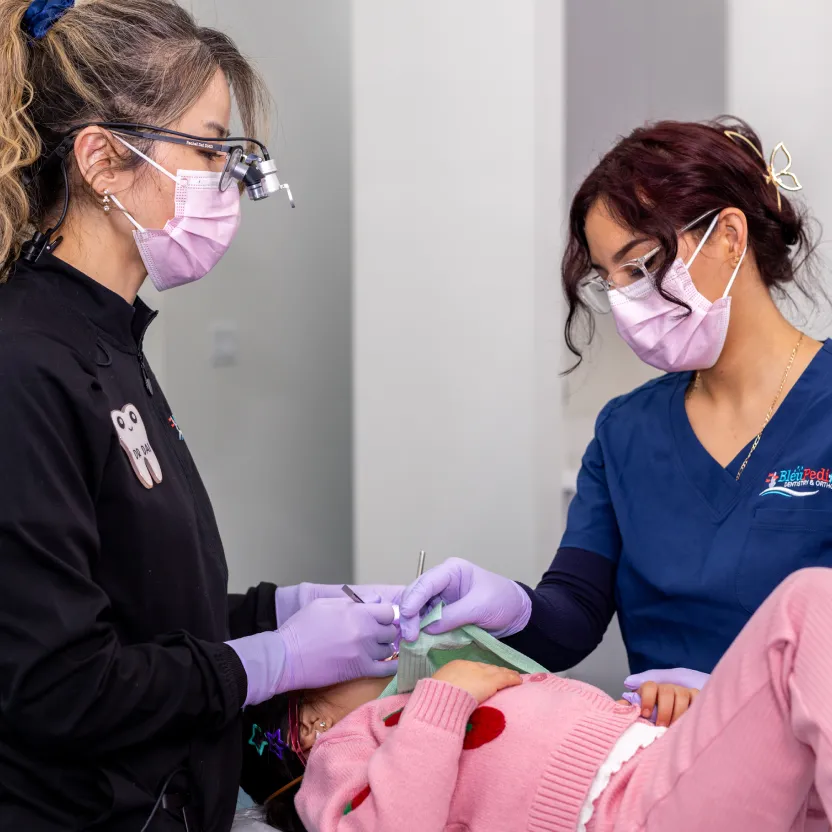 Two dental professionals wearing masks and gloves treating a young child lying in a dental chair.