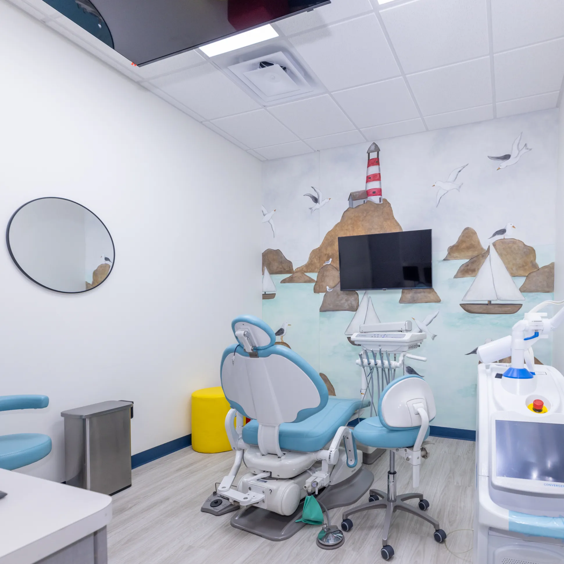 Clean dental examination room with blue dental chair, a mural of lighthouse and sailboats, and medical equipment.
