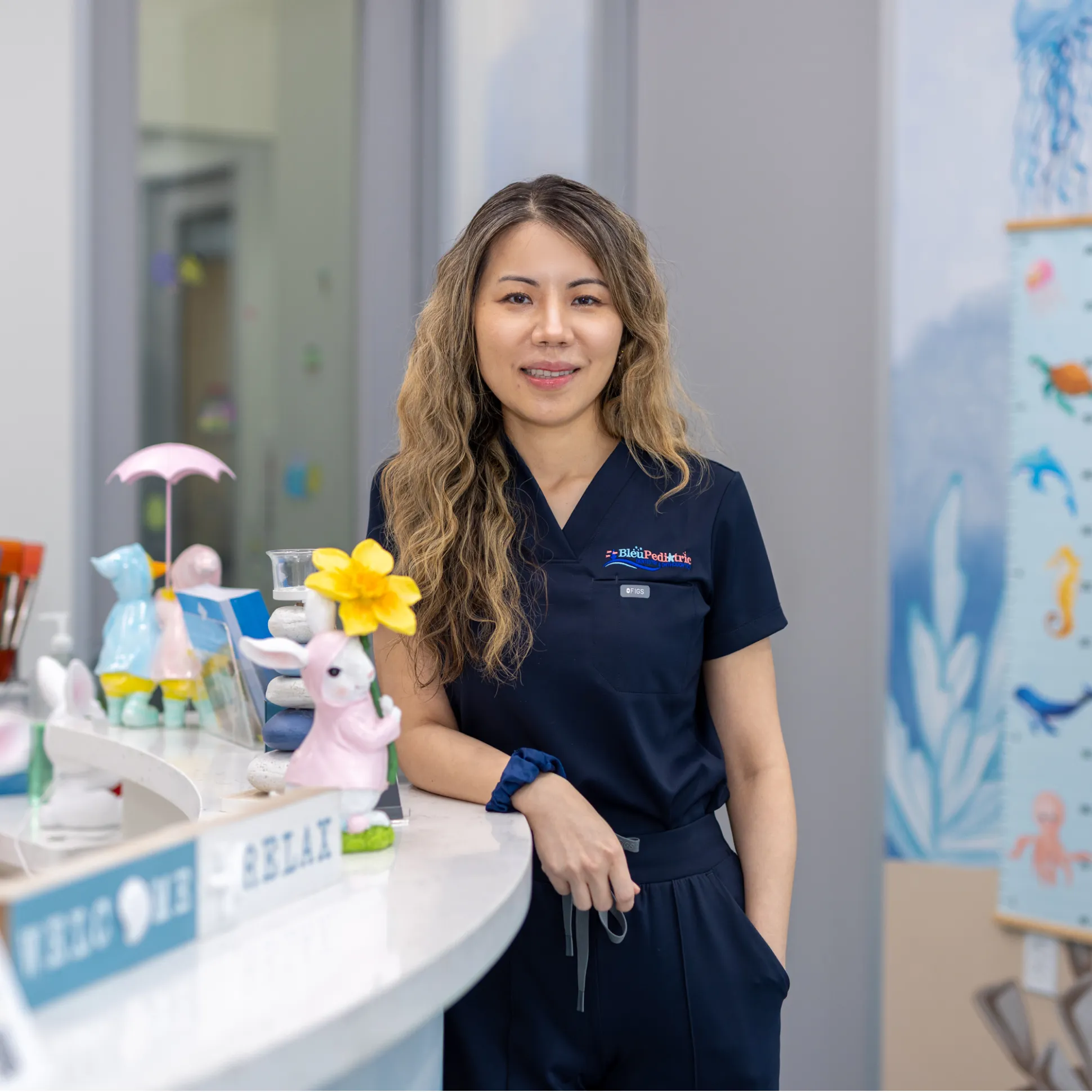 A smiling woman in navy scrubs leaning on a reception desk decorated with rabbit figurines and a yellow flower.