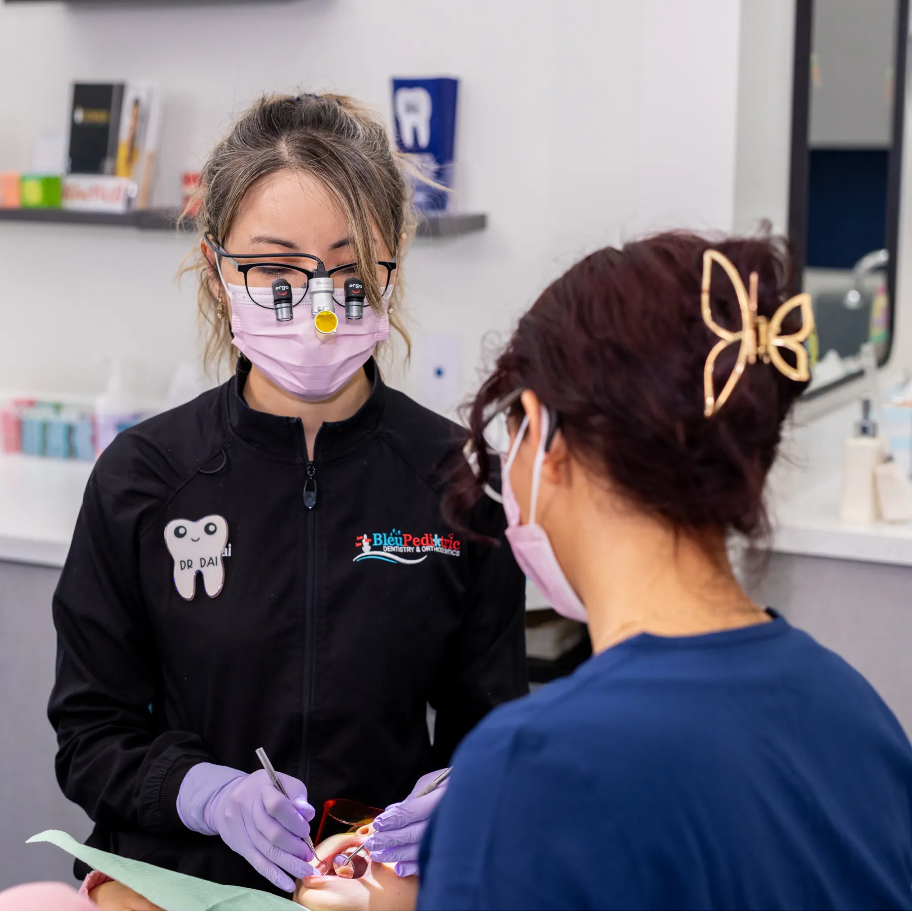 Dentist wearing magnifying loupes and a pink mask treating a young patient in a dental office while an assistant looks on.