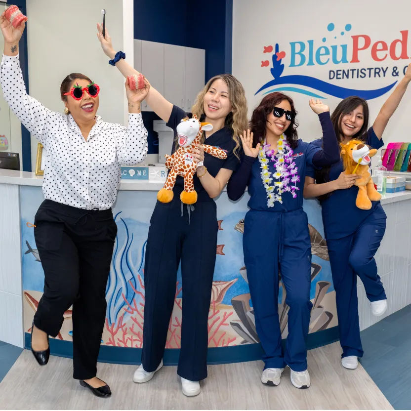 Four dental staff members in a pediatric dental office joyfully posing with dental models and stuffed animal toys in front of a reception desk with ocean-themed mural and Bleu Ped Dentistry logo.