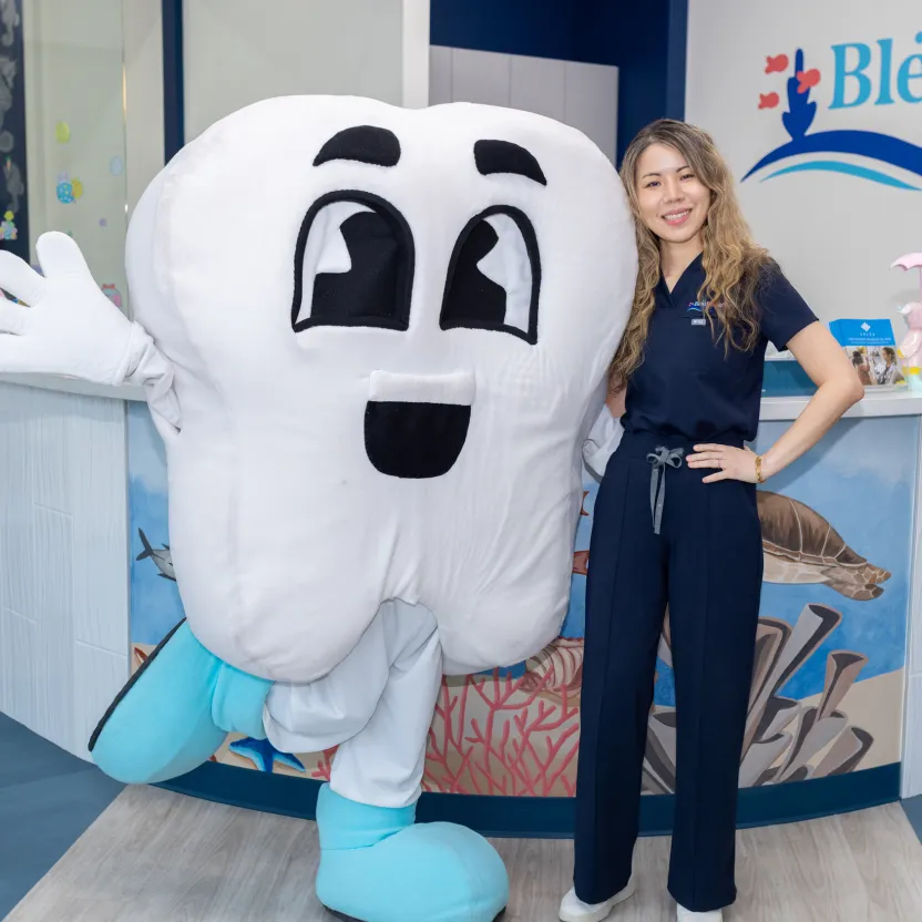 Woman standing with a large smiling tooth mascot wearing blue shoe covers in a dental office.
