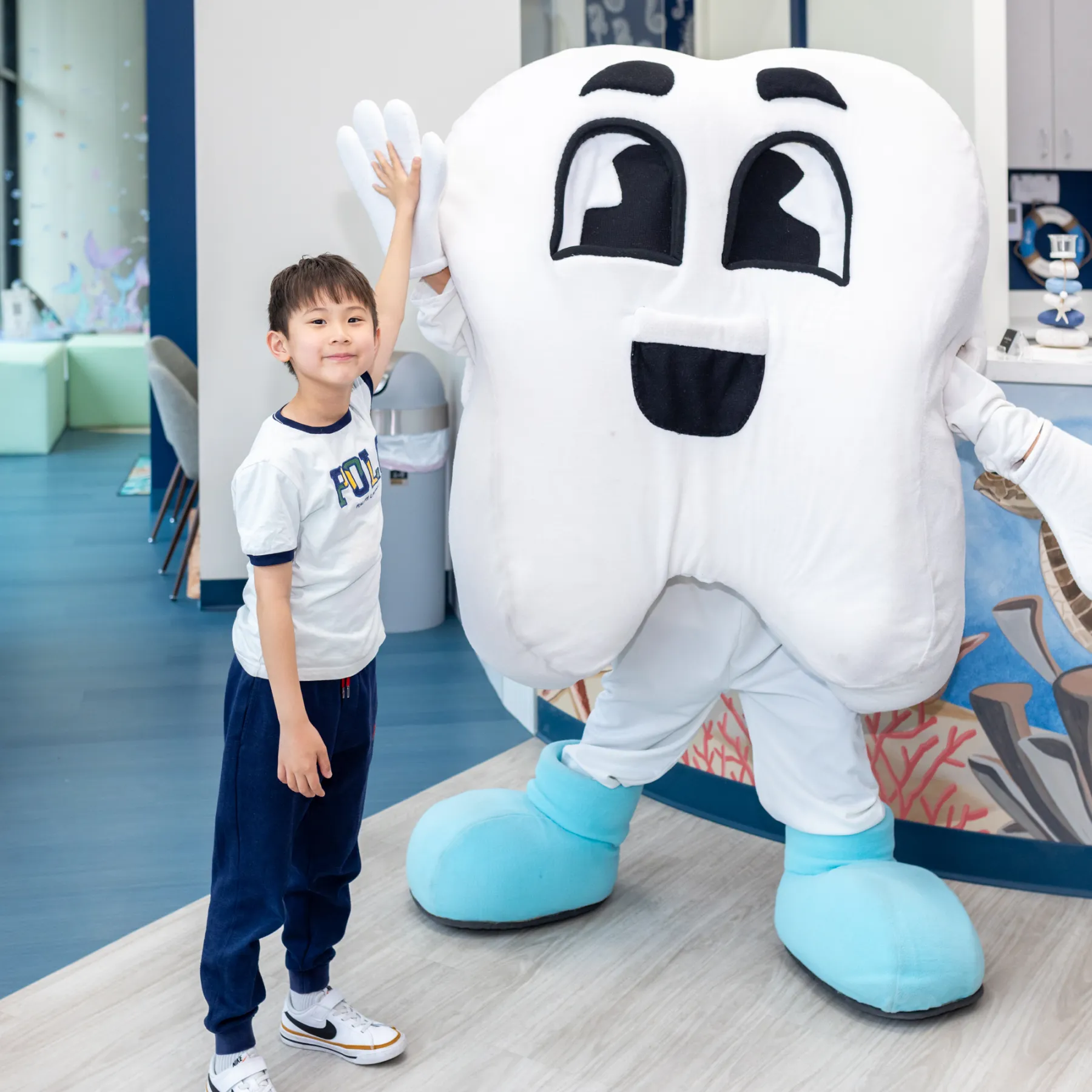 Young boy giving a high five to a person in a large smiling tooth mascot costume with blue shoes inside a dental office.