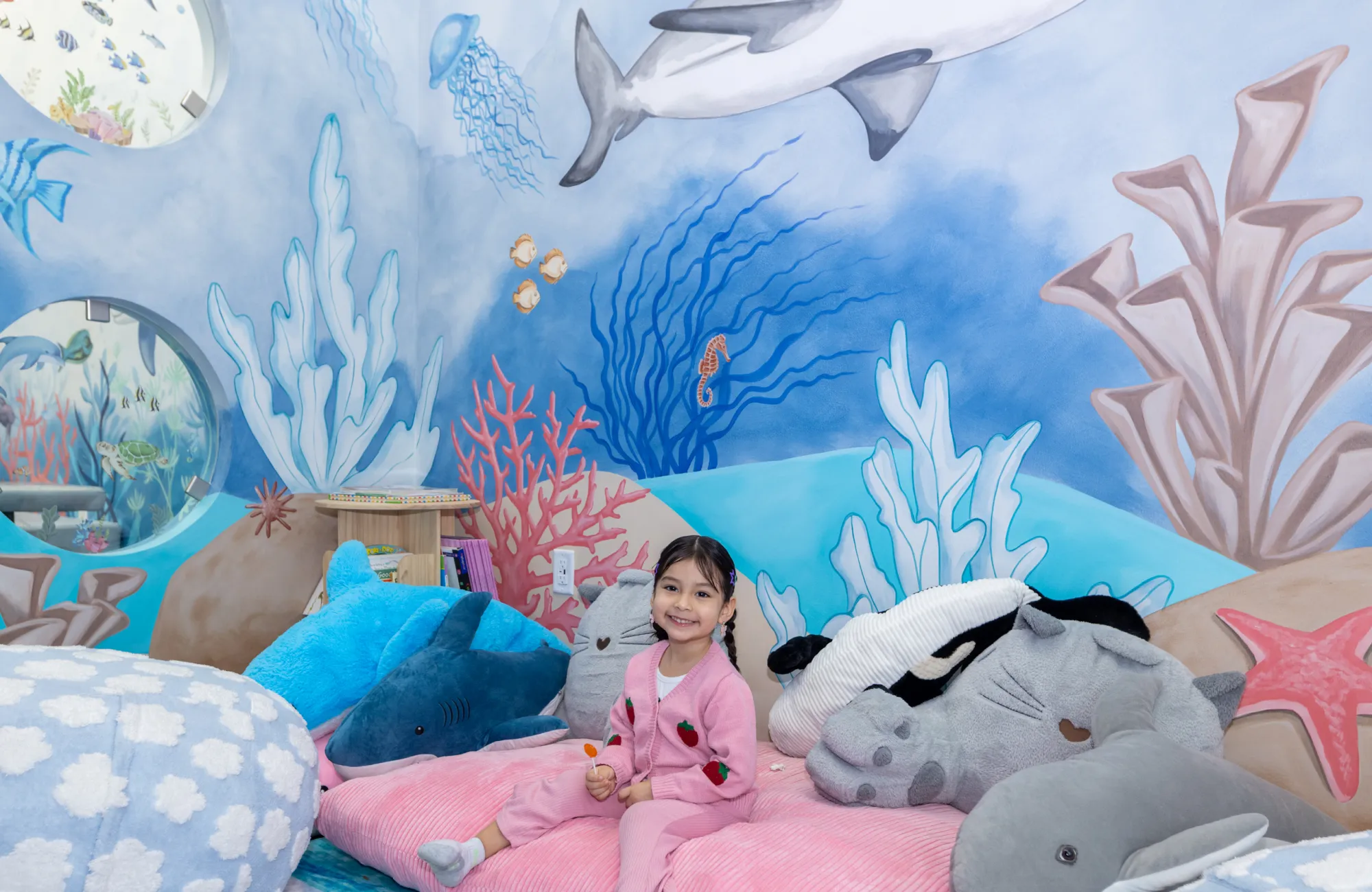 Smiling young girl in pink sitting on a bed with ocean animal plush toys in a room decorated with an underwater mural.
