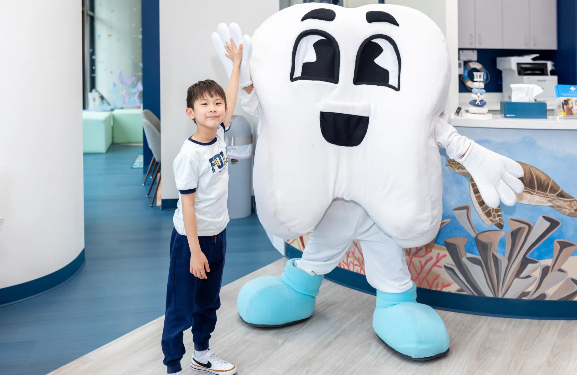 Young boy giving a high-five to a person in a large tooth mascot costume inside a dental office.