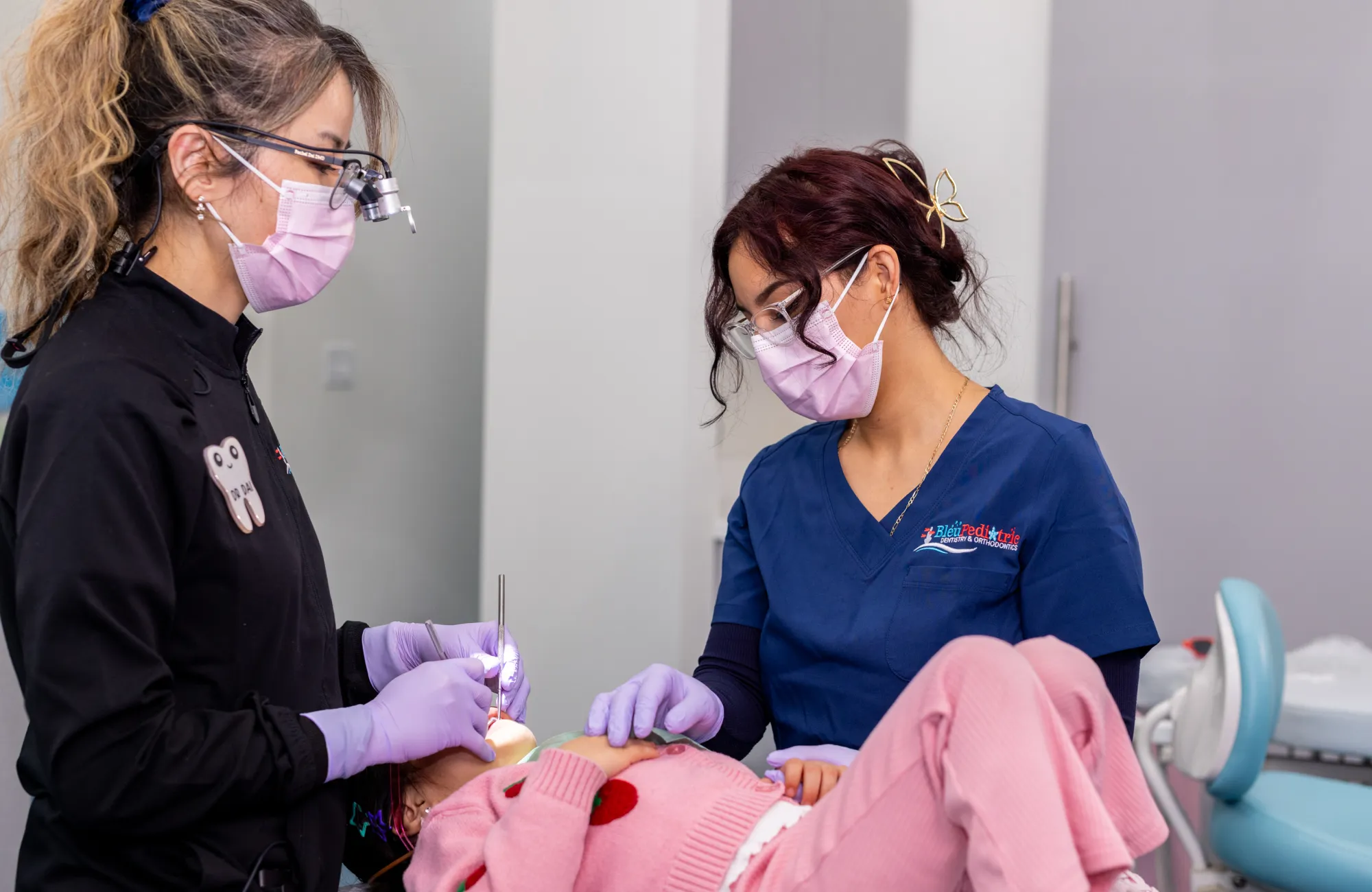 Two female dental professionals wearing masks and gloves examining a child patient lying in a dental chair.
