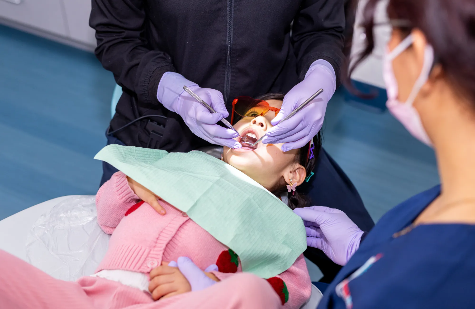 Young girl in pink sweater receiving dental checkup from a dentist wearing purple gloves and protective glasses, with an assistant nearby.